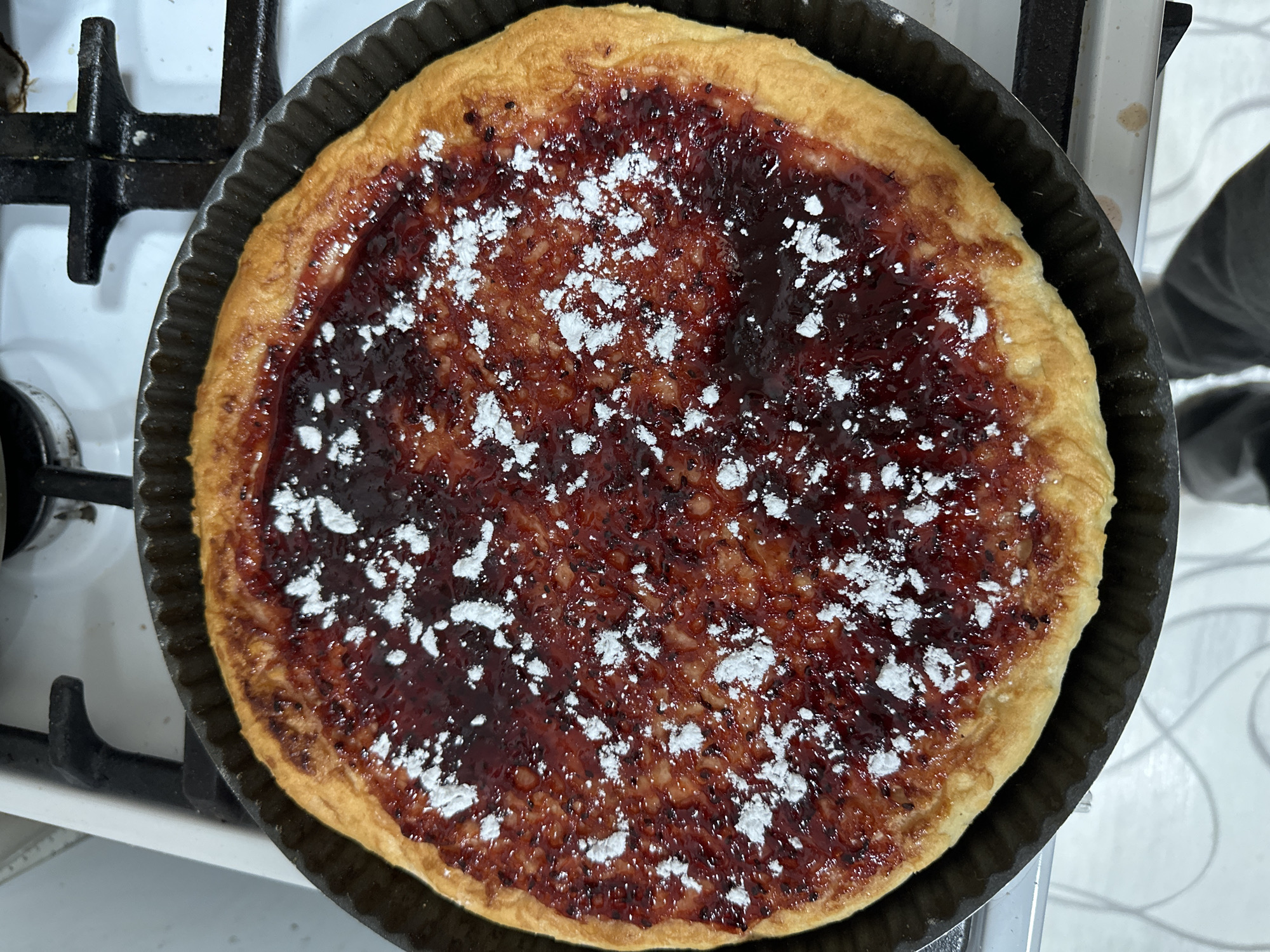 A pie with a red jam-like filling and powdered sugar is presented in a round, fluted baking pan. The pie rests on a gas stovetop. The location is not discernible as being in Сосново, Russia, based on visual information.