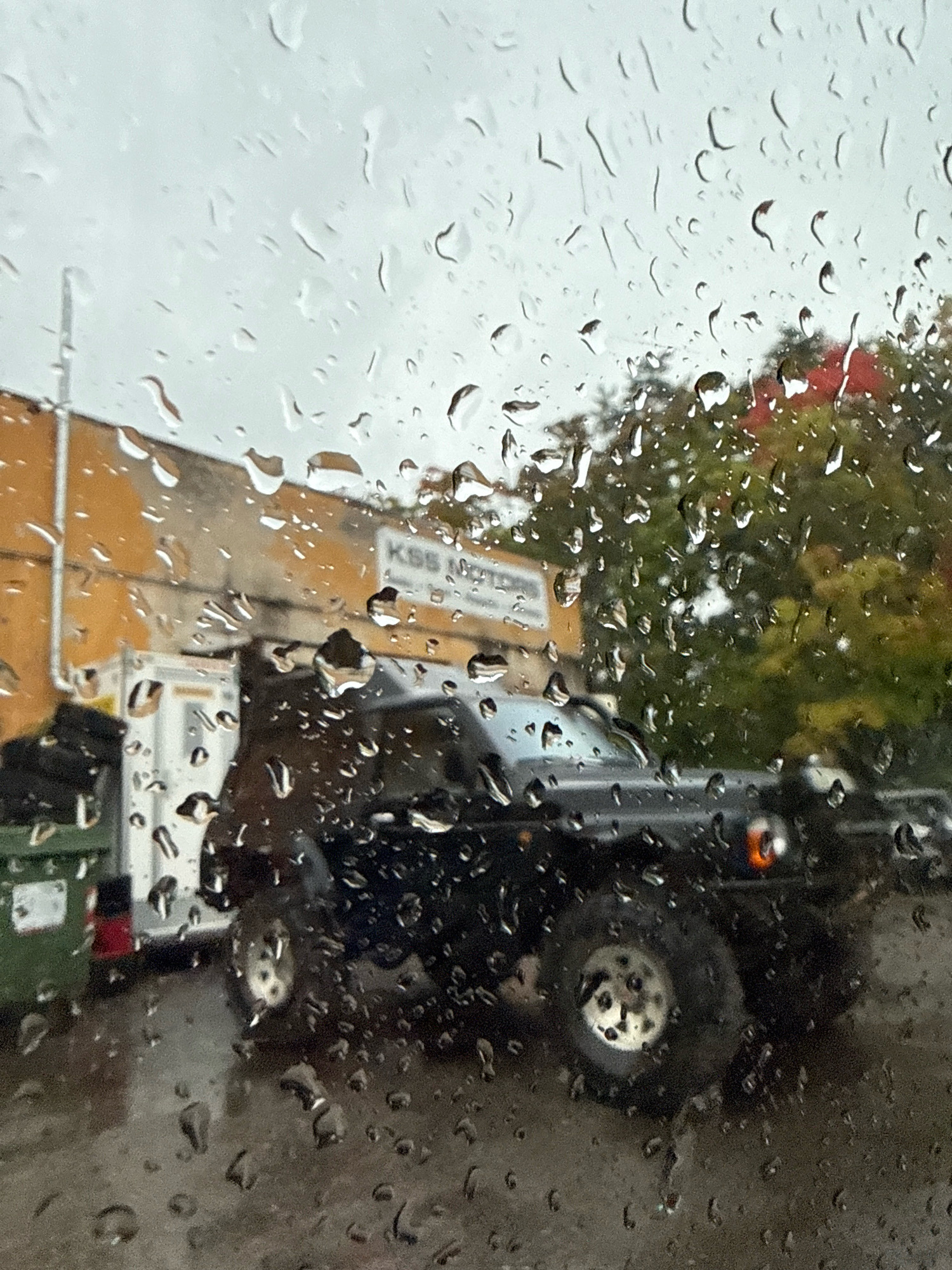 This is a medium shot, eye-level, slightly angled, exterior, daytime photograph taken through a rain-streaked window. The scene appears to be an industrial or commercial area, possibly a parking lot or service bay, with a focus on a dark-colored SUV.
The foreground is dominated by numerous water droplets clinging to the window, distorting the view of the background. These droplets vary in size and shape, with some appearing as streaks and others as distinct spheres.
In the middle ground, a black SUV is parked. Its tires are visible, with one large wheel prominent in the lower right quadrant of the image. The SUV's headlights are partially visible. Behind the SUV, a white building with an orange facade is partially obscured by rain. A sign on the orange building reads "KSS Motors". To the left of the SUV, a large green dumpster is visible, with part of a white structure or trailer behind it.
The background consists of trees with green and some reddish-brown foliage, suggesting an autumn or late summer setting. The sky is overcast and grey, contributing to the overall damp atmosphere.
No individuals are clearly visible in the photograph. The image conveys a sense of a rainy, overcast day in an urban environment. The specific location in Tallinn, Estonia, cannot be determined from the visual information alone.