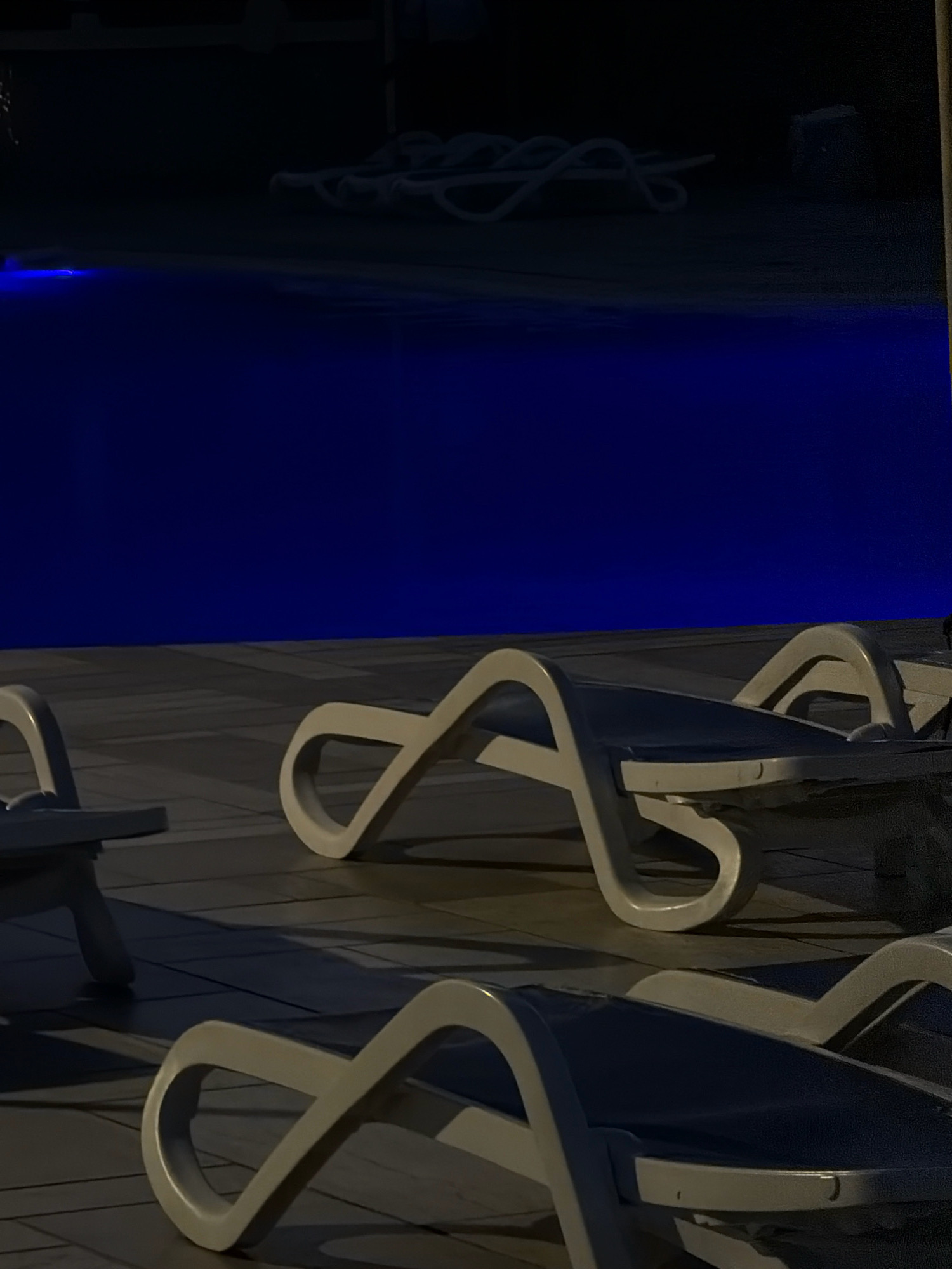The image depicts an outdoor scene at night, likely a resort or hotel area, with several white lounge chairs positioned on a tiled patio next to a swimming pool. The pool is illuminated with a deep blue light, creating a vibrant contrast with the dark surroundings. The lounge chairs are made of white plastic and have a modern, curved design. Some of the chairs are empty, while others appear to have dark blue fabric seats. Shadows are cast across the patio, indicating the direction of unseen light sources. In the background, a coiled white rope or cable is visible on the patio, and the far end of the area is shrouded in darkness. No people are present in the image. The scene suggests a tranquil and inviting atmosphere, ideal for relaxation by the pool. Given the context of Sharm El Sheikh, Egypt, this could be a common amenity found in many of the city's tourist establishments, particularly along the Red Sea coast.