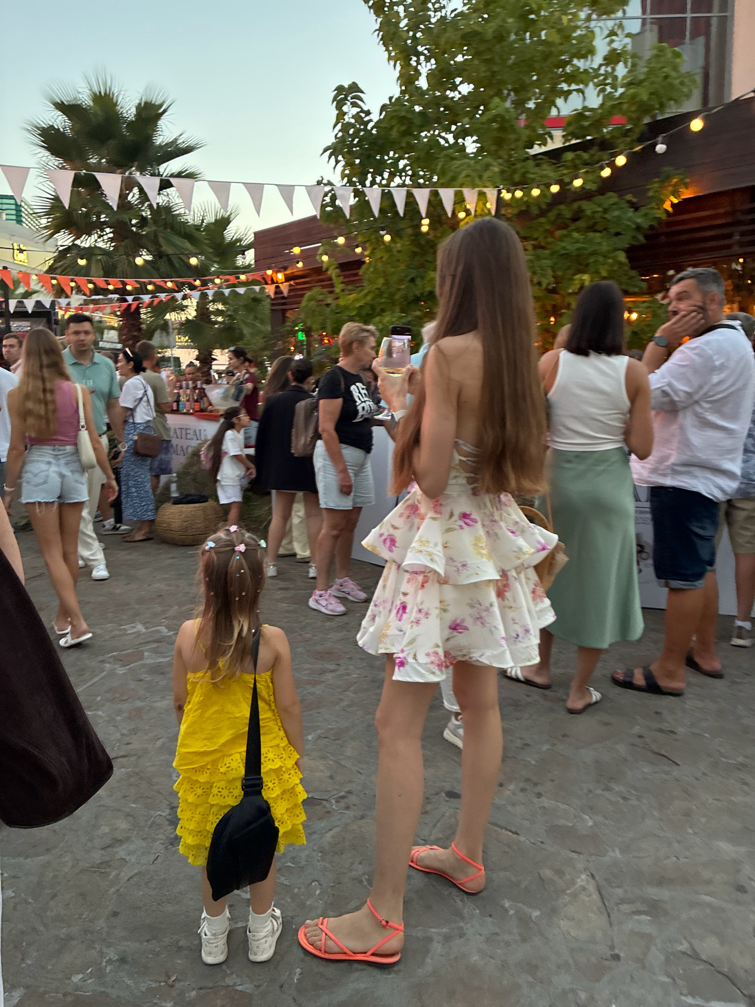 In Sochi, Russia, an outdoor event is underway, possibly a festival or market, with string lights and triangular flags decorating the scene. In the foreground, a young girl in a bright yellow, tiered dress and white socks with sandals stands facing away from the camera, wearing a black bag slung over her shoulder. To her right, a tall woman in a floral, tiered dress and bright orange sandals is also facing away. Other individuals, including men and women in casual summer attire, are visible in the background, some interacting with each other, while others are walking or standing. Palm trees and lush greenery are interspersed with structures, suggesting a warm, possibly coastal, environment. The overall atmosphere appears relaxed and festive.