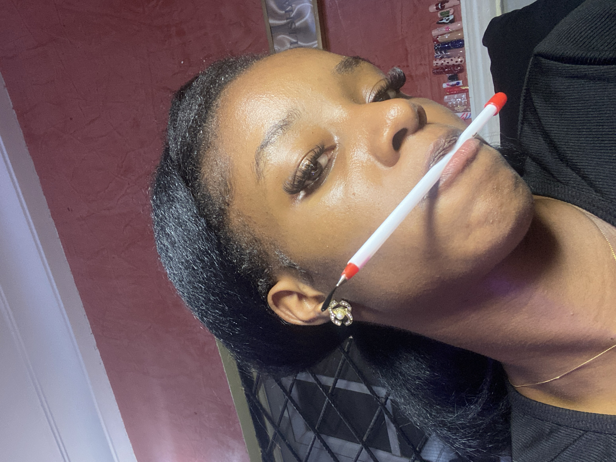 A medium close-up shot shows a young Black woman with dark hair, wearing a black top and a gold necklace. Her face is turned to the side, and she is holding a white pen with a red tip horizontally in her mouth. She has on false eyelashes and a prominent earring on her left ear. Behind her, a wall painted a deep red is visible. To her right, a shelf displays a collection of decorative artificial nails. The overall lighting is somewhat dim, suggesting an indoor setting, possibly in Jos, Nigeria.