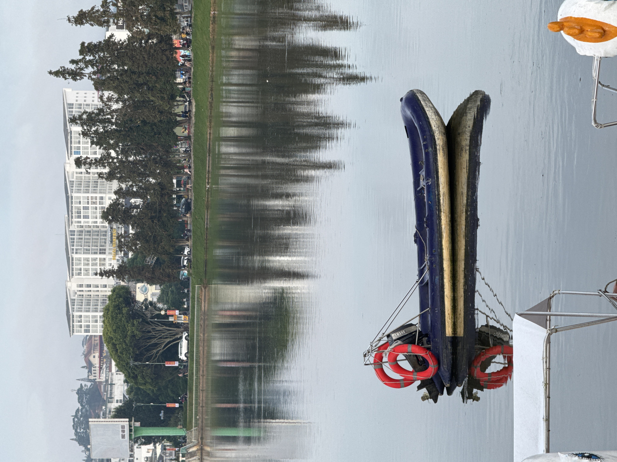 The image depicts a serene view of Xuan Huong Lake in Da Lat, Vietnam. In the foreground, a dark blue inflatable boat with two red life preservers is anchored in the calm water, its reflection clearly visible. To the left and right of the inflatable boat, parts of other boats are visible, one with a white canopy and another with a white and orange life preserver. The middle ground features a grassy embankment leading to a tree-lined promenade with several people and parked vehicles. Dominating the background are modern white buildings and lush green trees, interspersed with some older structures. The sky is overcast, contributing to the muted tones of the scene.