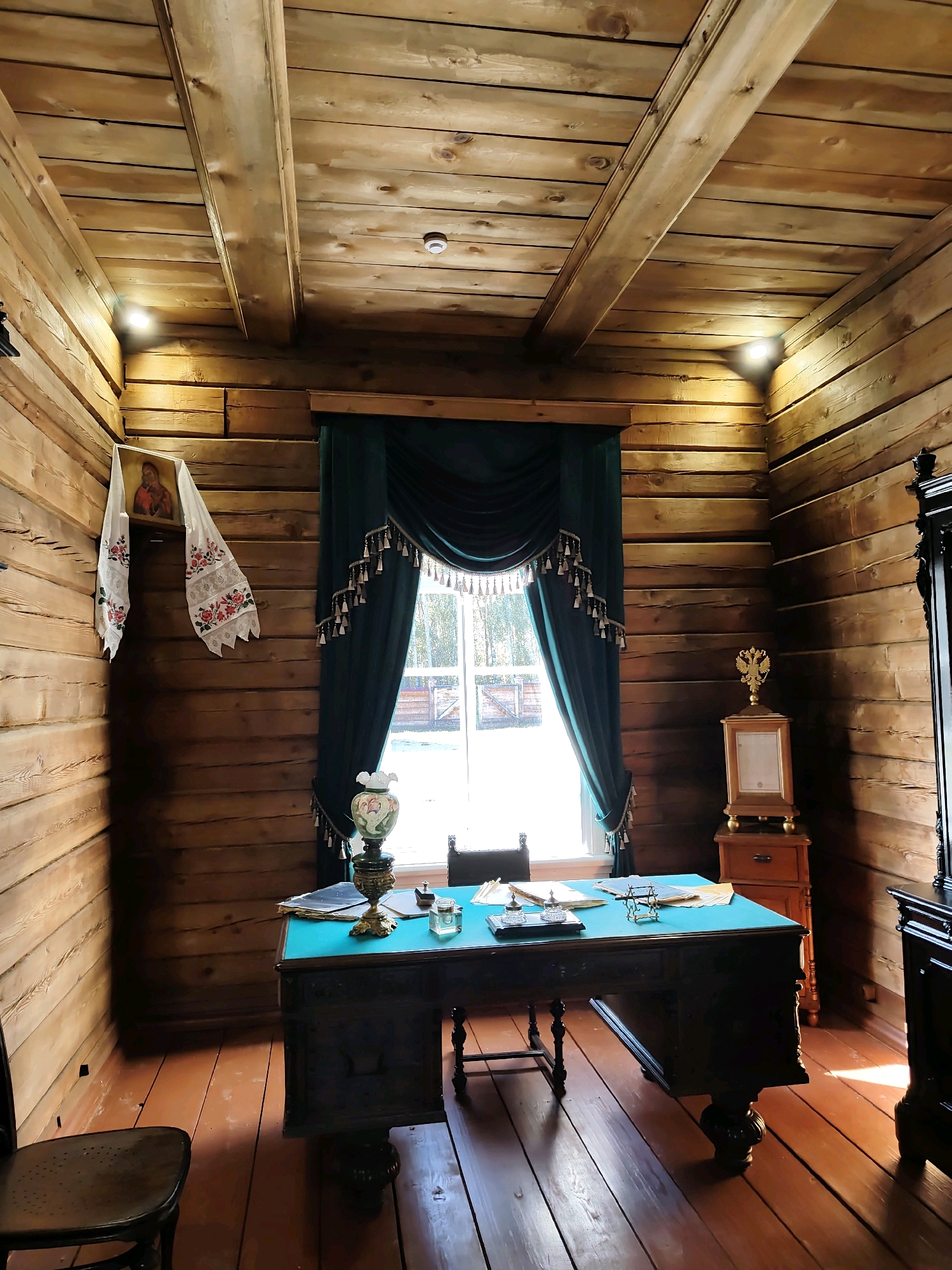 The image depicts an interior scene of a room with wooden walls and a wooden beamed ceiling, likely a historical setting in the Taltsy (Irkutsk), Russia museum. In the center of the room, a dark wooden desk with a teal-colored surface is positioned in front of a window. The desk is adorned with a vintage lamp, papers, a small inkwell, and a decorative item. A dark green curtain with tassels frames the window, which offers a glimpse of an outdoor scene with vegetation and a wooden fence. To the left of the window, a framed icon of a religious figure is hung on the wall, accompanied by a white embroidered textile. To the right of the window, a wooden cabinet with a drawer sits beneath a framed document topped by a double-headed eagle emblem. A dark wooden chair is placed to the left of the desk, and another chair is partially visible to the right. The floor is made of polished wooden planks, reflecting the sunlight streaming through the window. The overall impression is of a preserved historical office or study.