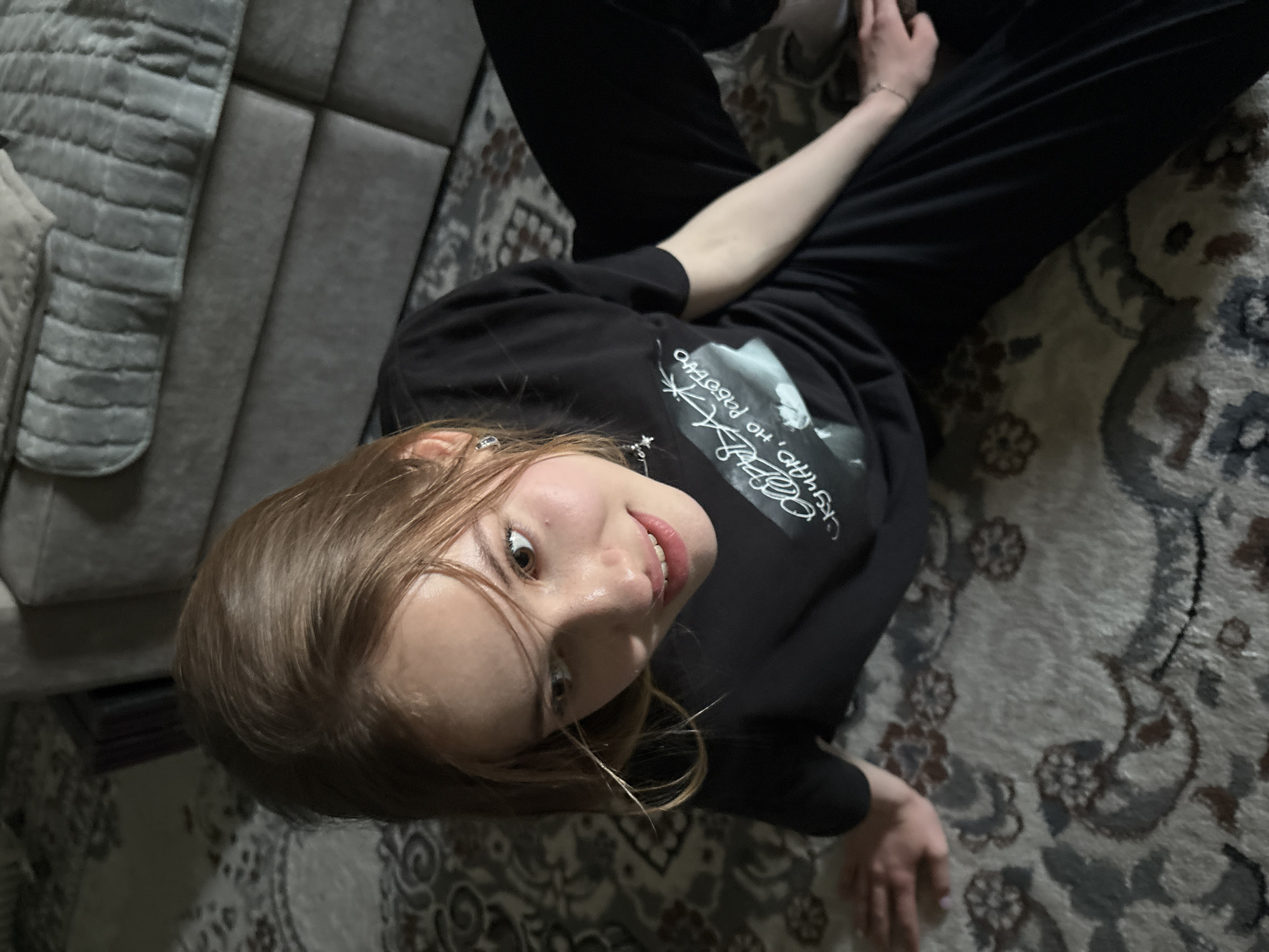 A young person with long, light brown hair and wide, light-colored eyes is lying on their back on a rug. They are wearing a black t-shirt with white text and graphics, and black pants. Their head is tilted back, and they are looking directly at the camera with a slightly surprised expression, a smile on their lips. Their right arm is bent and resting on their chest, while their left arm is extended. The background shows a grey, tufted couch or headboard on the left and a patterned rug covering the floor. The lighting appears to be artificial and dim. There is white text visible on the t-shirt, which seems to be in Cyrillic script. The scene suggests a casual, indoor setting, possibly a home. The time of day and weather are not discernible.