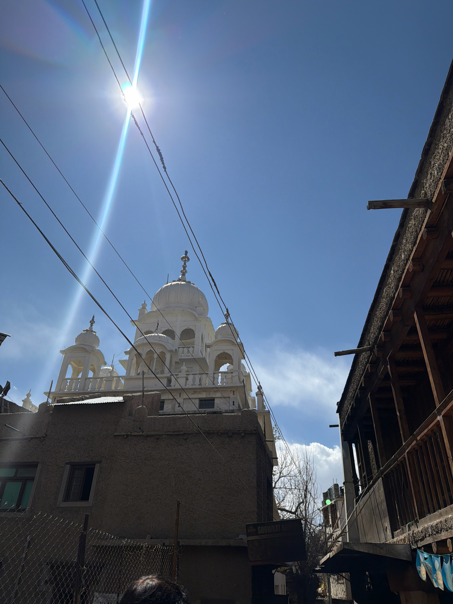 This is a low-angle, outdoor, daytime photograph featuring a white domed building, likely a Gurudwara, in Leh, India. The building is adorned with multiple smaller domes and intricate architectural details. In the foreground, a rough-textured building with a tin roof is visible, and a wire fence separates it from the viewer. To the right, a wooden structure with a balcony and exposed beams is prominently featured. The sky is a clear blue with a few scattered clouds, and the sun is intensely bright, creating lens flare and a lens flare effect across the image. Power lines are strung across the upper portion of the frame, obscuring parts of the sky and the white domed building. The overall scene suggests a street view in an urban or semi-urban environment, with a mix of traditional and utilitarian architecture.