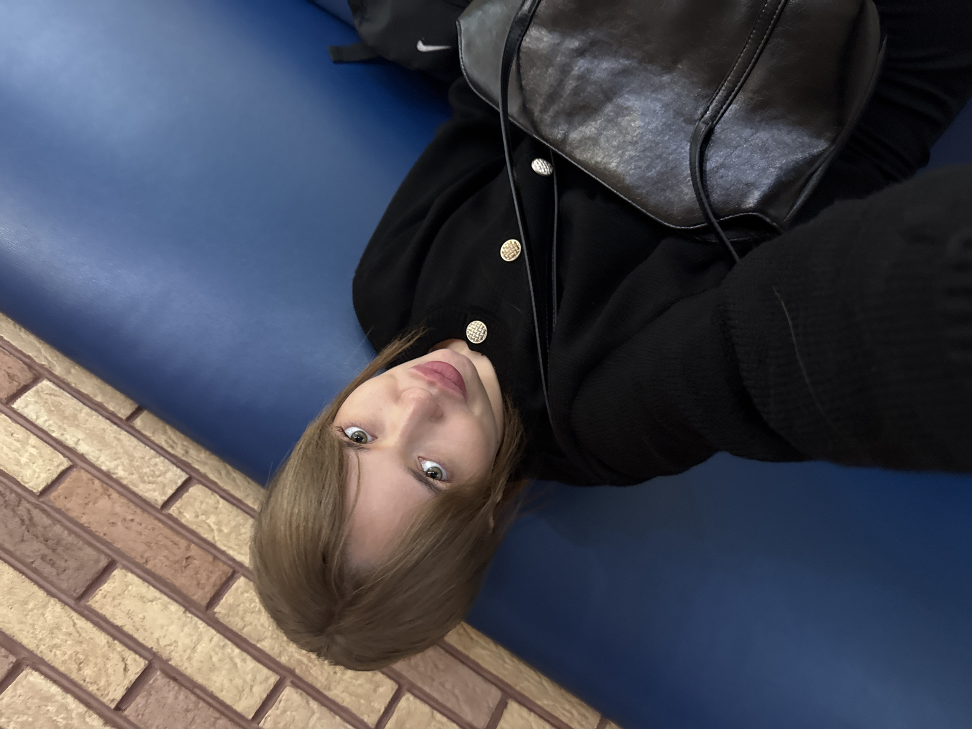 A woman with blonde hair is lying on a blue surface. She is wearing a black sweater with gold buttons and a black bag. The surface she is lying on is next to a brick wall. Based on the presence of the distinctive blue color and brick texture, this could potentially be a location in the city of Chaikovsky, Russia, near a building with a blue-painted exterior and a brick wall. The woman is looking at the camera. A portion of a black backpack is visible in the upper left corner of the frame.