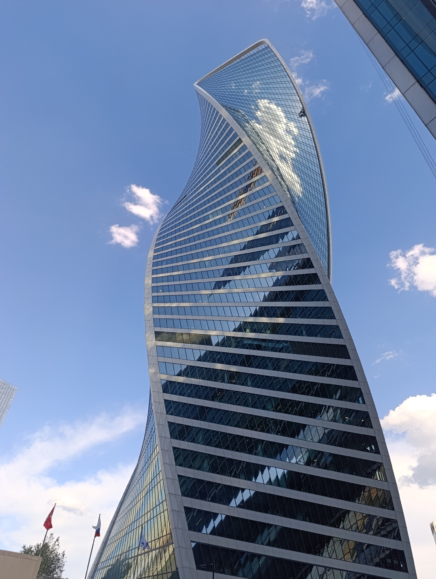 A dynamic low-angle shot captures the distinctive twisted design of a modern skyscraper under a bright blue sky with scattered white clouds. The building, identified as the Evolution Tower in Moscow, Russia, features a helical shape with its glass and metal facade appearing to rotate as it ascends.
The skyscraper's exterior is predominantly composed of reflective glass panels, which mirror the sky, clouds, and the surrounding urban environment, creating abstract patterns and distortions. Horizontal bands of light grey or white material separate the window sections. High up on the building's curved glass facade, a small figure, likely a window cleaner, is visible, engaged in maintenance work.
To the far left of the main building, another glass-fronted structure is partially visible. On the bottom left of the frame, three flags on poles are displayed: a solid red flag, the white-blue-red tricolor flag of Russia, and a light blue flag with a white emblem. Some green foliage from trees can be seen near the base of the flags. To the far right, a sliver of another tall, modern building is also visible, hinting at a dense urban setting.
The time of day appears to be sunny daytime, with clear visibility and a few fluffy clouds. The overall impression is one of modern architecture within a bustling city landscape on a pleasant day. There is no visible text in the image.