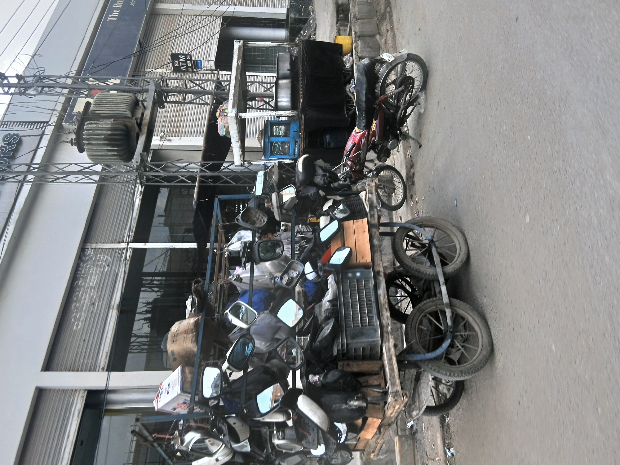The image shows a street scene in Quetta City Tehsil, Pakistan. A cart loaded with motorcycle parts, including mirrors, seats, and tires, is parked along the roadside. A motorcycle is also parked nearby. The buildings in the background appear to have commercial storefronts with closed metal shutters. Overhead, there's a utility pole with a transformer. The street is paved and appears to be relatively busy.