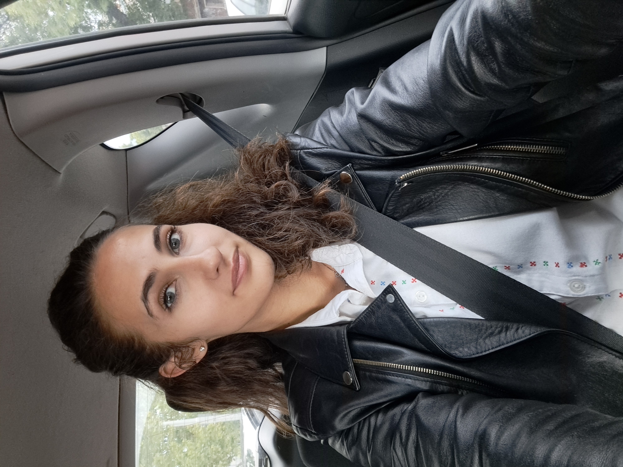 The image is a selfie taken inside a car. A young woman with curly brown hair and light eyes is wearing a black leather jacket over a white shirt with small colored floral patterns. She is wearing a seatbelt. The interior of the car is visible, including parts of the dashboard and door. The background outside the car shows trees and possibly a building. It is not possible to confirm the exact location within Sofia, Bulgaria, based on the information in the image.