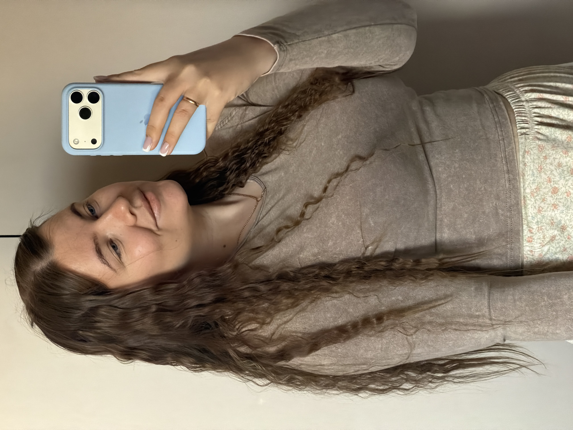 A young woman is taking a selfie indoors. She is holding a blue iPhone with a distinctive triple-lens camera design. She is wearing a long-sleeved, light brown top and floral-patterned pajama bottoms. She has long, curly brown hair. She is wearing a ring on her ring finger. The background appears to be a plain, light-colored wall and floor. There is no visual evidence to indicate the specific location of Chaikovsky, Russia.