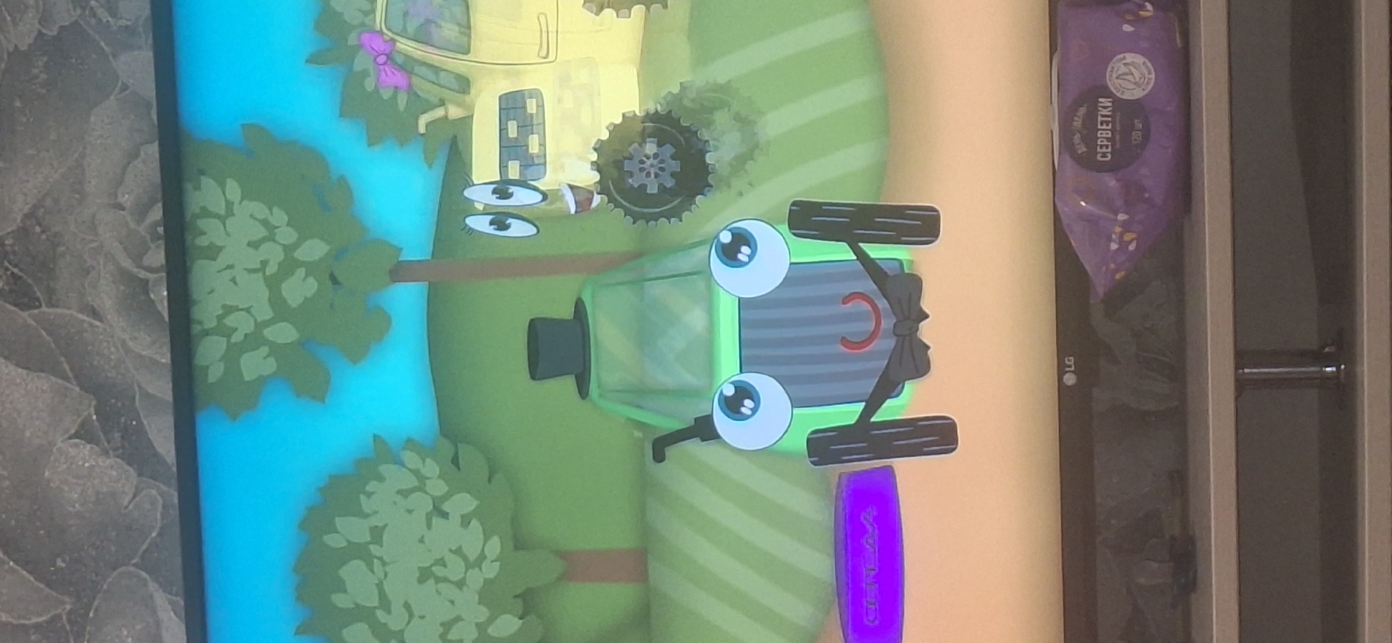 A cartoon image depicts two tractors in a grassy field with trees under a blue sky. The tractor in the foreground is green, has large blue eyes, a red crescent-shaped mouth, and wears a black top hat and a black bowtie. It has black wheels and a black exhaust pipe. To the left of the green tractor is a purple object with a white inscription that is difficult to read. The tractor in the background is yellow, has large eyes and eyelashes, and a pink bow on its roof. The scene is displayed on a television screen, with the bottom of the screen showing a shelf with a package of wet wipes and a visible "LG" logo on the television itself. The image is not located in the city of Bila Tserkva, Ukraine.