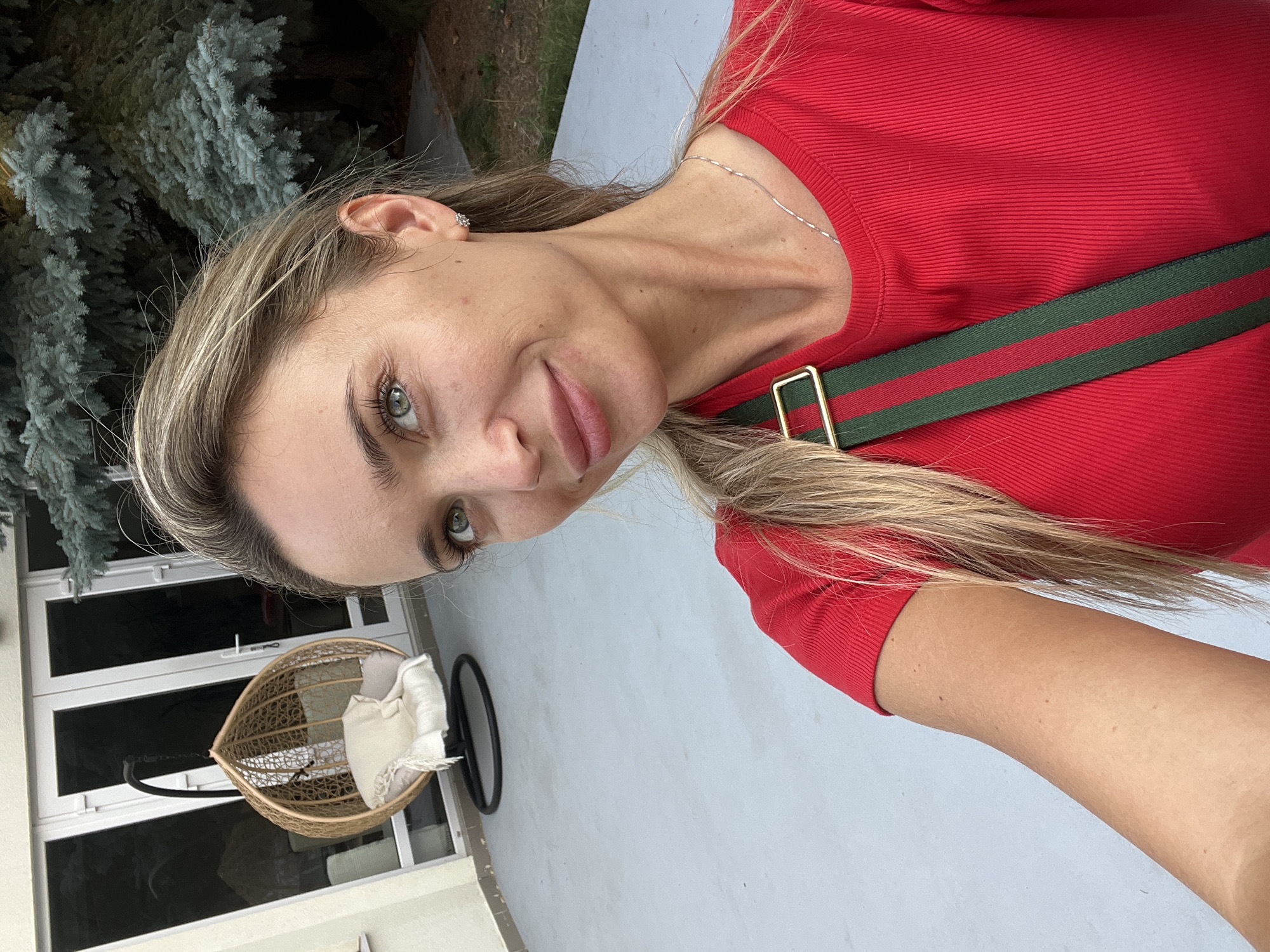 A medium-shot, outdoor selfie shows a blonde woman with green eyes, fair skin, and a light dusting of freckles. She is wearing a red, ribbed t-shirt and a green and red striped strap with a gold buckle crosses her chest. Her hair is pulled back into a ponytail. She is smiling and looking slightly off-camera to her left. In the background, a light gray patio is visible, along with a beige building with large glass doors and a hanging egg chair. To the right of the woman, a blue-green pine tree looms. The setting appears to be a private residence with an outdoor patio area. The image does not provide any information suggesting a location in Fontanka, Ukraine.