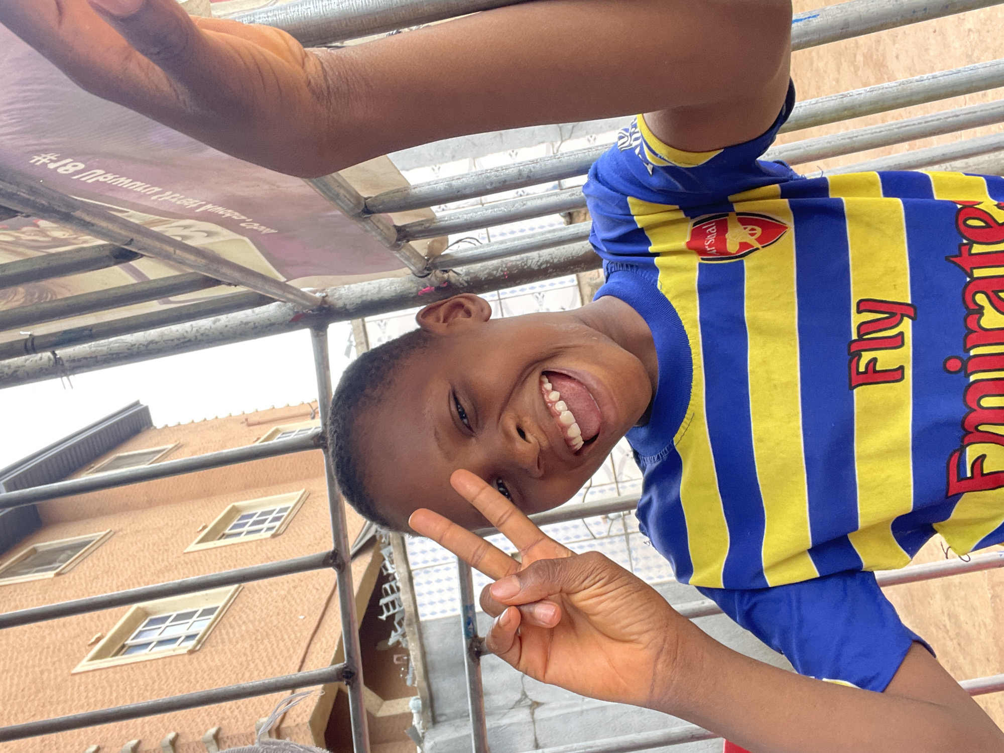 A young person in an Arsenal soccer jersey smiles broadly, flashing a peace sign with one hand. The shot is taken from a low angle, looking up through metal bars towards a multi-story peach-colored building with multiple windows, typical of architecture in Onitsha, Nigeria. Overhead, more metal bars and what appears to be a promotional banner are visible. The young person is partially upside down in the frame.
