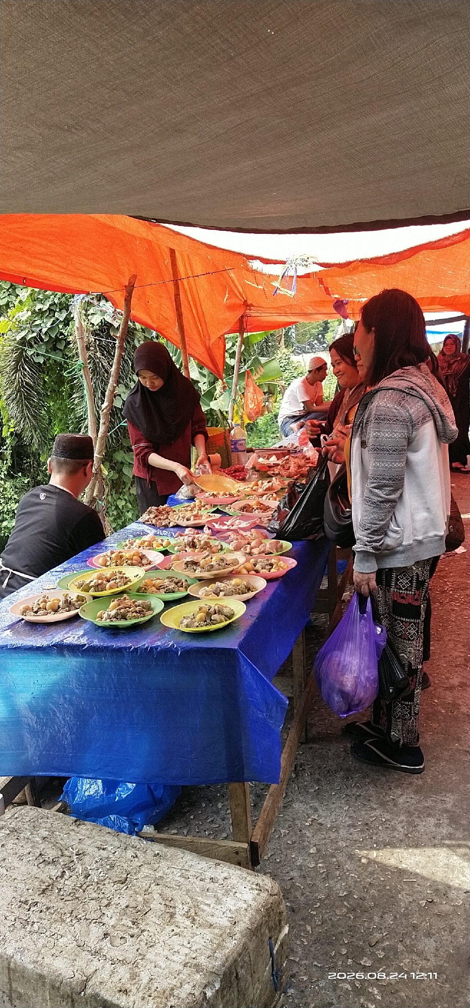 The image depicts an outdoor market scene in Tamiang Layang, Indonesia, likely a street food stall. An orange tarp serves as a canopy, supported by wooden poles, providing shade. Beneath the canopy, a long blue-covered table is laden with numerous small plates of food, predominantly featuring what appear to be small pieces of meat and eggs.
Several individuals are present. A woman wearing a black hijab and a maroon shirt stands behind the table, appearing to serve food. To her left, a man in a black t-shirt and a black peci hat is seated, with his back to the camera. To the right of the serving woman, two women are engaged in conversation, one in a gray patterned hoodie and the other in a yellow top. Further in the background, a man in a white shirt and white cap is seated. Another woman in a dark hijab is visible in the far right background.
The ground is rough, textured concrete or asphalt. The overall impression is of a lively, informal marketplace offering prepared food. The date stamp "2026.08.24 12:11" is visible in the lower right corner.