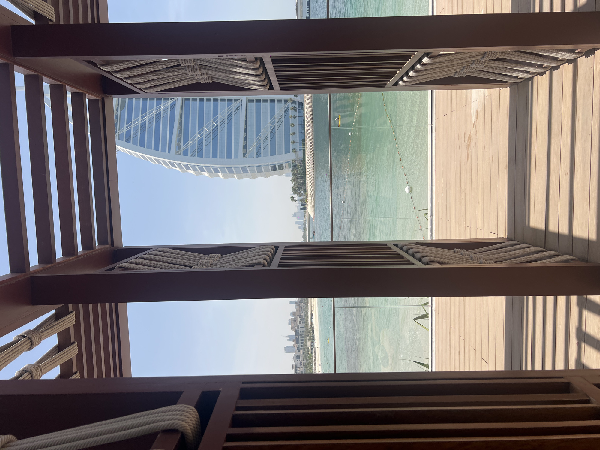 The image shows a framed view of the Burj Al Arab hotel in Dubai, United Arab Emirates. The framing structure is made of dark brown wood, with slats and thick ropes woven in a decorative pattern. The view extends beyond the hotel to the sea, where a lifeguard line is visible, with a wooden deck on one side. In the background, there is also a view of the Dubai skyline.