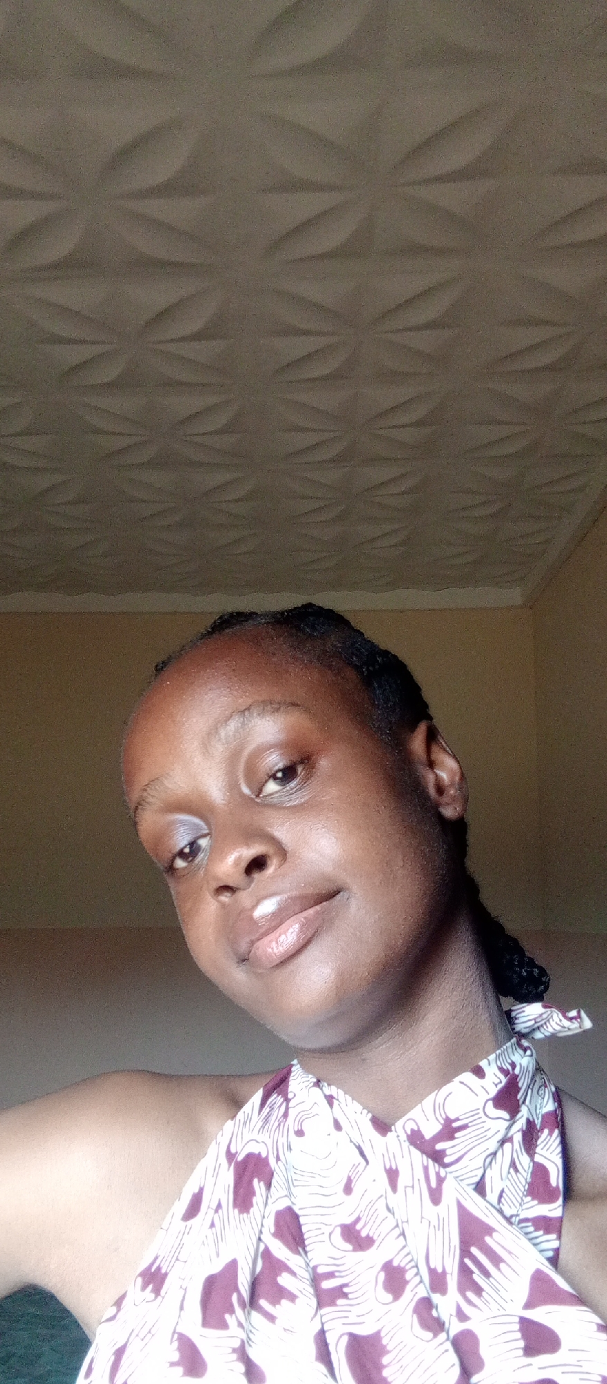 A close-up portrait features a young Black woman looking directly at the camera with a gentle, slight smile. Her head is tilted slightly to her right. She has dark skin with a natural sheen, braided hair pulled back, and appears to be wearing light makeup including a subtle shimmery eyeshadow on her left eyelid and clear lip gloss.
She is wearing a patterned garment, possibly a halter neck or wrap top, which is white with abstract burgundy or maroon shapes that resemble stylized hands or organic forms.
The background consists of a plain, light brown wall on the right side and a textured ceiling above. The ceiling features a repeating three-dimensional pattern of interlocking leaf-like or floral motifs. The lighting is bright and appears to be natural daylight, illuminating her face evenly. There is no visible text in the image. The setting is indoors, with the provided context being Akwanga, Nigeria.