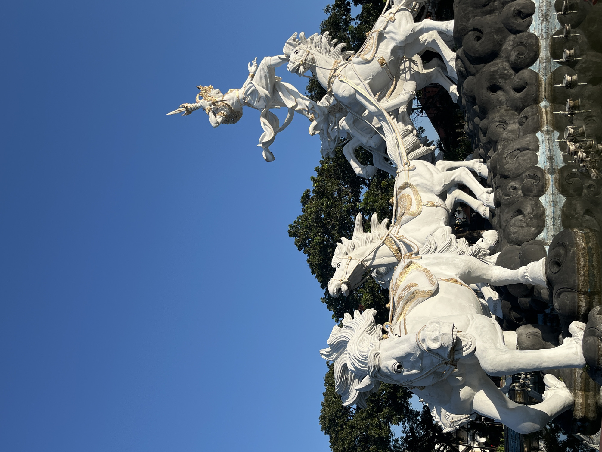 This image captures a white sculpture of a deity riding a chariot pulled by horses against a clear blue sky. The sculpture appears to be made of white stone or plaster, with decorative gold accents on the horses' harnesses and the deity's crown and garment. The deity is depicted in a dynamic pose, leaning forward with a spear-like object in one hand, suggesting movement and action. The horses are also depicted in mid-stride, with their manes flowing. The sculpture is partially obscured by dark, textured elements that resemble clouds or rock formations, and a lush green tree can be seen behind the horses. The lighting suggests it is daytime, likely morning or afternoon, with bright sunlight illuminating the sculpture. The overall scene conveys a sense of power and majesty. Based on the location context of Kuta, Indonesia, this sculpture is likely part of a larger monument or fountain, possibly at a temple or public space. There is no visible text in the image.