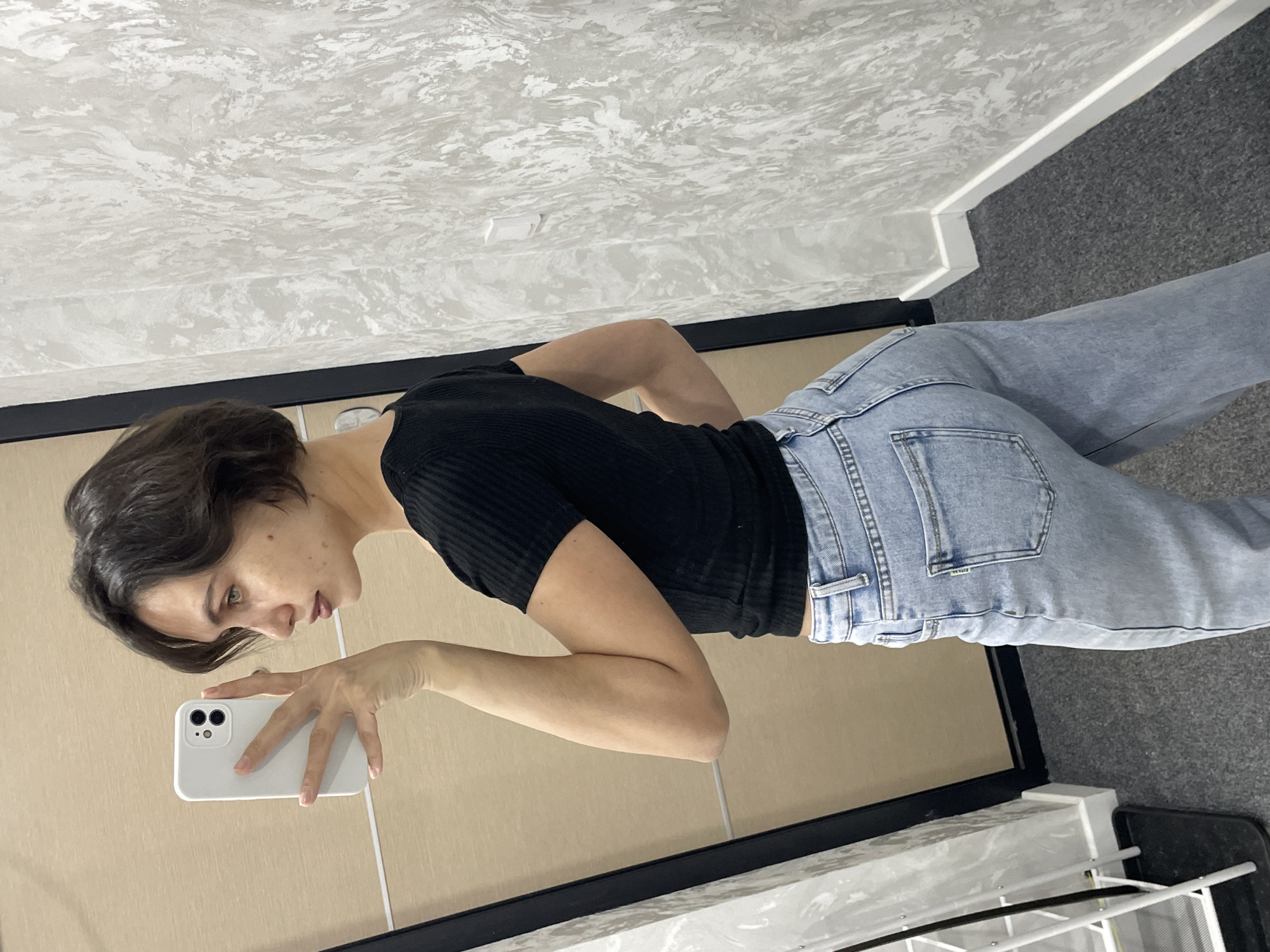 A young woman with short brown hair is taking a selfie in a bathroom. She is wearing a black short-sleeved top and light blue jeans. She is holding a white smartphone in her left hand. The bathroom has a white and grey patterned wall, a tan-colored wall, and a grey tiled floor. A black frame is visible in the foreground. The location in Izhevsk, Russia is not discernible from the image.