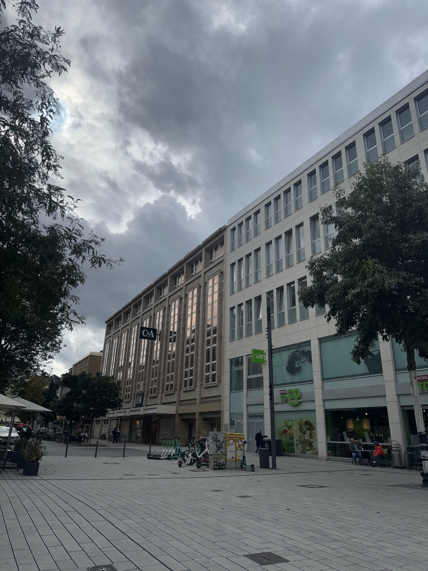The image depicts a street scene in Dusseldorf, Germany, under a heavily overcast sky. The view is predominantly of a modern multi-story building with a light-colored facade and numerous windows. To the left, an older building with a beige facade and tall, narrow windows is visible, featuring a prominent "C&A" logo. The street in the foreground is paved with gray rectangular stones, and several parked electric scooters are lined up. Further down the street, a store with a green "REWE GO" logo is open, with some customers seated outside. Trees with dark green foliage are present on both sides of the street, with a larger tree to the right of the frame. Several individuals are visible, some walking, others sitting at tables. The overall atmosphere is subdued, consistent with a cloudy day.