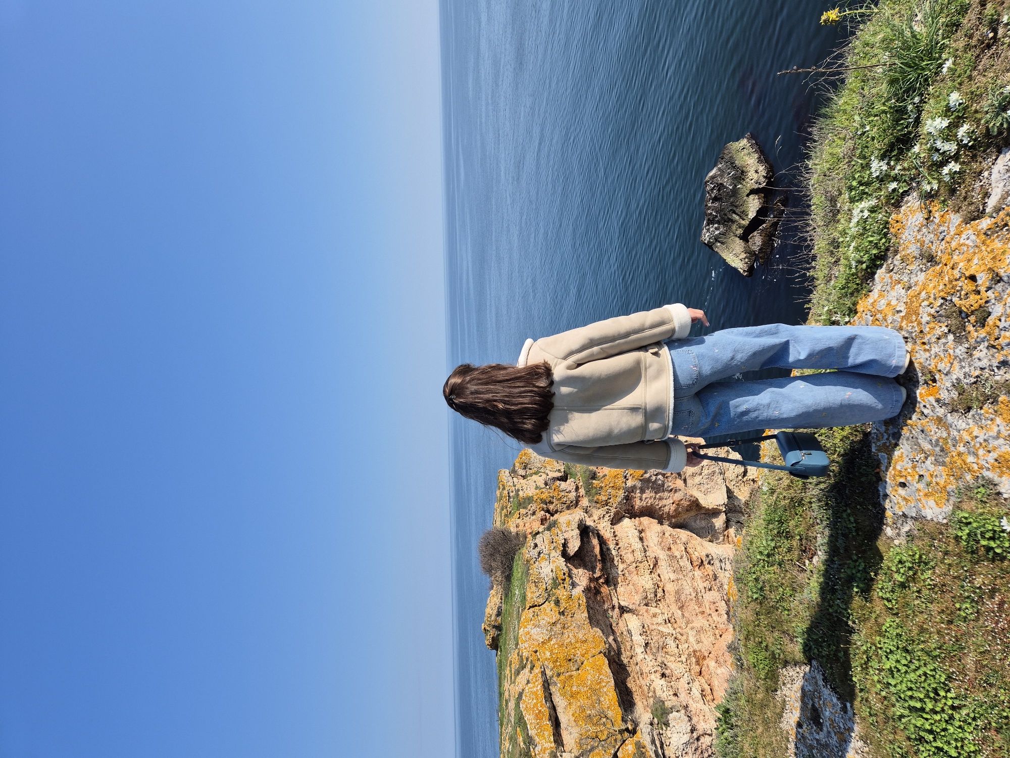 A young woman stands on a cliff overlooking the Black Sea near Kamen Bryag, Bulgaria. The woman is seen from the back, with long brown hair, wearing a cream-colored jacket and blue jeans. The cliff's edge is covered in green grass and some white and yellow flowers, with orange lichen growing on the rock face. Below the cliff, the dark blue sea meets a rocky outcrop. Above, the clear blue sky extends towards the horizon. The woman is also carrying a blue bag.