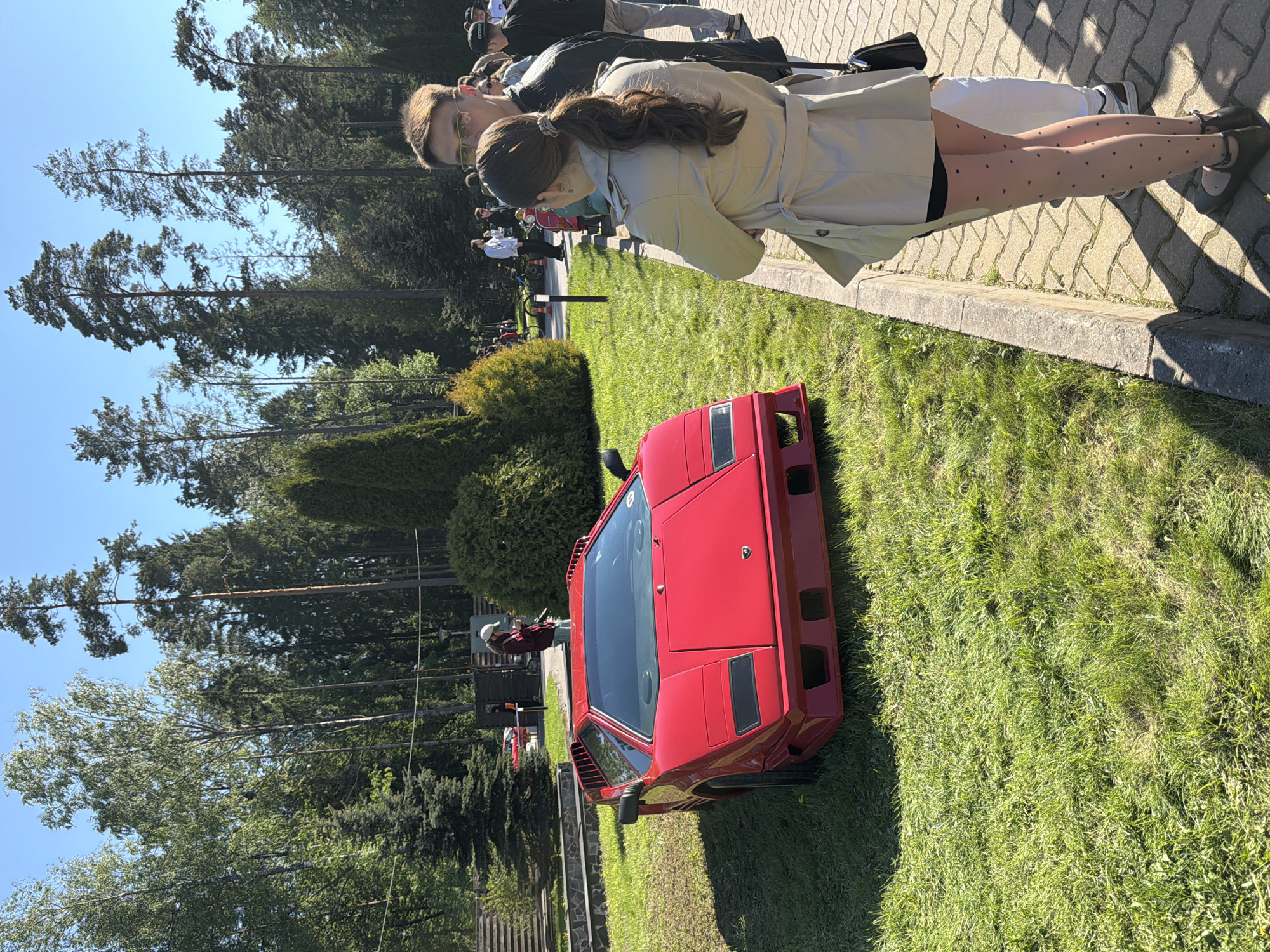The image shows a red Lamborghini Countach parked on grass at an outdoor car show in Repino, Russia. Two people, a man and a woman, are standing near the car, looking at it. The woman is wearing a light-colored coat and tights with black polka dots, while the man is wearing glasses. There are other people and cars visible in the background. Tall trees are also in the background. A paved walkway runs parallel to the lawn.