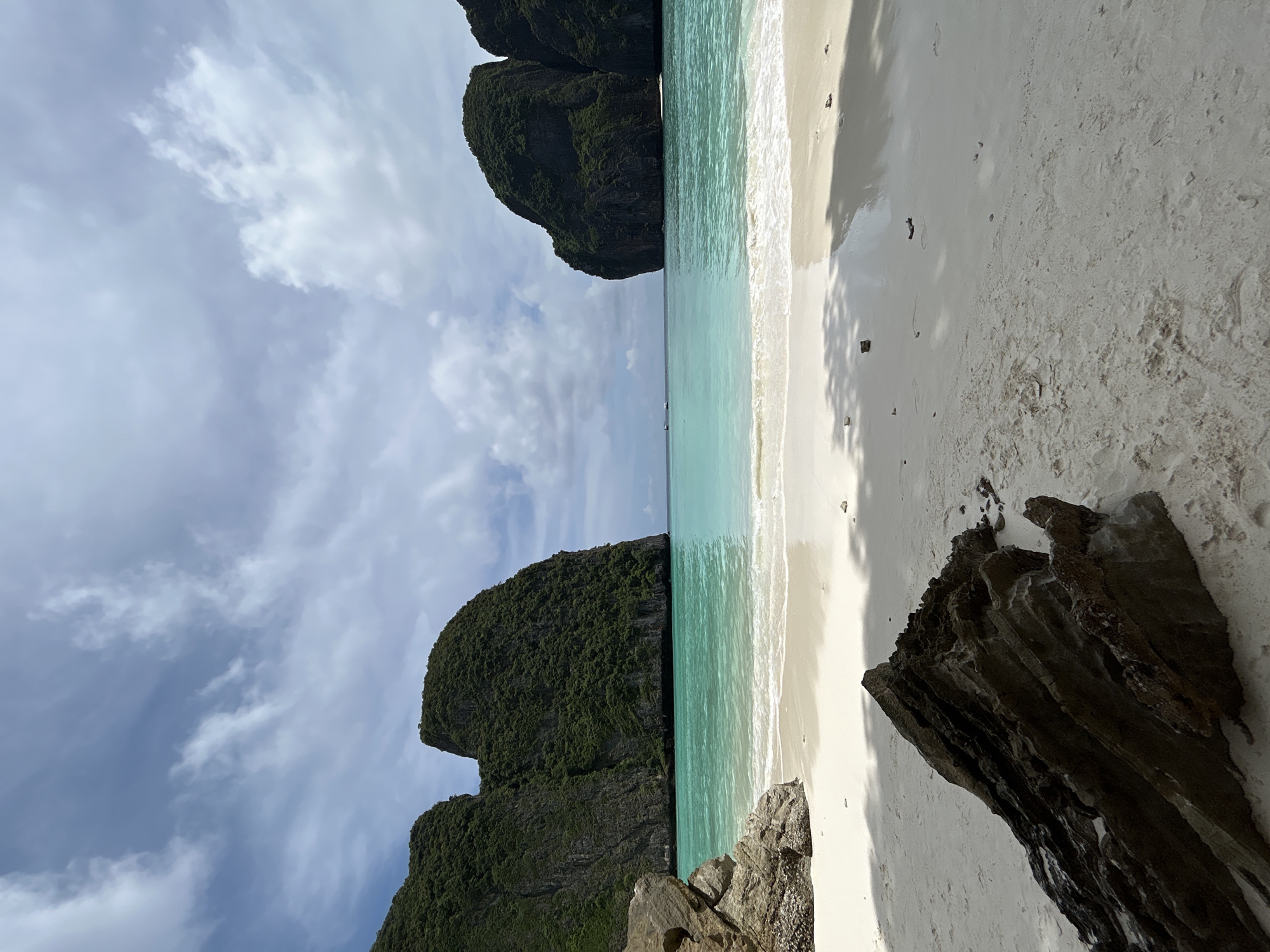 This image depicts Maya Bay, a stunning bay located on Koh Phi Phi Leh island in the Andaman Sea, Krabi Province, Thailand, near the city of Ao Nang. The scene is a tranquil tropical beach with powdery white sand and crystal-clear turquoise waters. Two large, lush green limestone karsts rise dramatically from the sea, flanking the bay. The water is calm and shallow near the shore, with gentle waves lapping at the beach. The sky is partly cloudy, with patches of blue sky peeking through scattered white clouds, suggesting fair weather. In the foreground, a large, dark, jagged rock formation rests on the sand, adding a touch of ruggedness to the otherwise idyllic landscape. Shadows from unseen foliage cast dappled patterns on the wet sand near the water's edge. Several small boats are visible in the distance on the water, indicating the presence of other visitors, though no people are directly visible in the frame. The overall atmosphere is serene and picturesque, highlighting the natural beauty of the location.