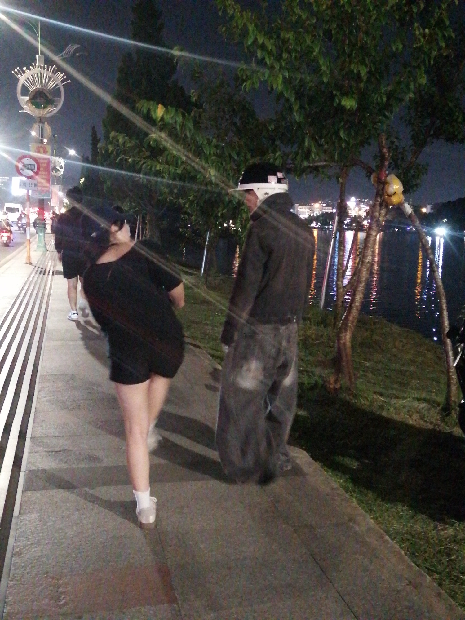 A nighttime street scene in Da Lat, Vietnam, shows two individuals on a sidewalk adjacent to a lake. The person in the foreground, facing away from the camera and towards the lake, is a female wearing a black dress and white socks with sandals. She is walking on the sidewalk. To her left, a male in a dark jacket, gray pants, and a helmet is walking towards the right of the frame, also on the sidewalk. In the background, a body of water reflects city lights, and trees line the shore. Streetlights illuminate the scene, creating lens flares. Traffic lights and a decorative street lamp are visible on the left side of the image.