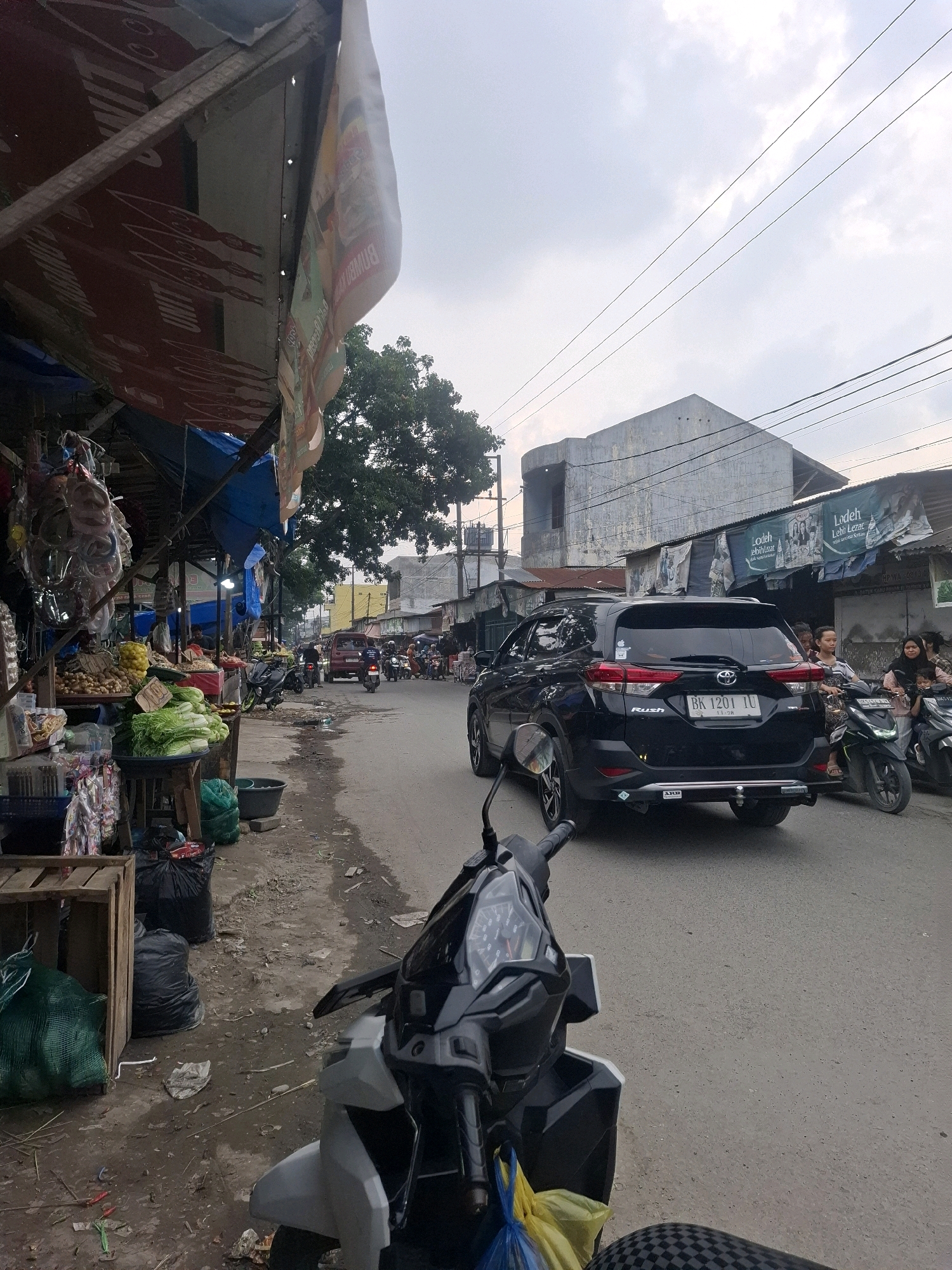 A street market scene is captured from the perspective of someone on a motorcycle. The market stalls, adorned with blue and red tarps, are on the left side of the road, displaying produce such as leafy green vegetables and what appear to be potatoes and lemons. Bags of produce and trash are scattered on the ground near the stalls. A black Toyota Rush SUV is driving on the right side of the asphalt road, followed by several motorcycles. A family is visible on one of the motorcycles. The street is lined with buildings, some with advertisements and weathered facades. Overhead, power lines crisscross the sky, which is overcast with clouds. The overall impression is a bustling, everyday scene in a market area within a city. The specific location is identified as Kenangan, Indonesia.
