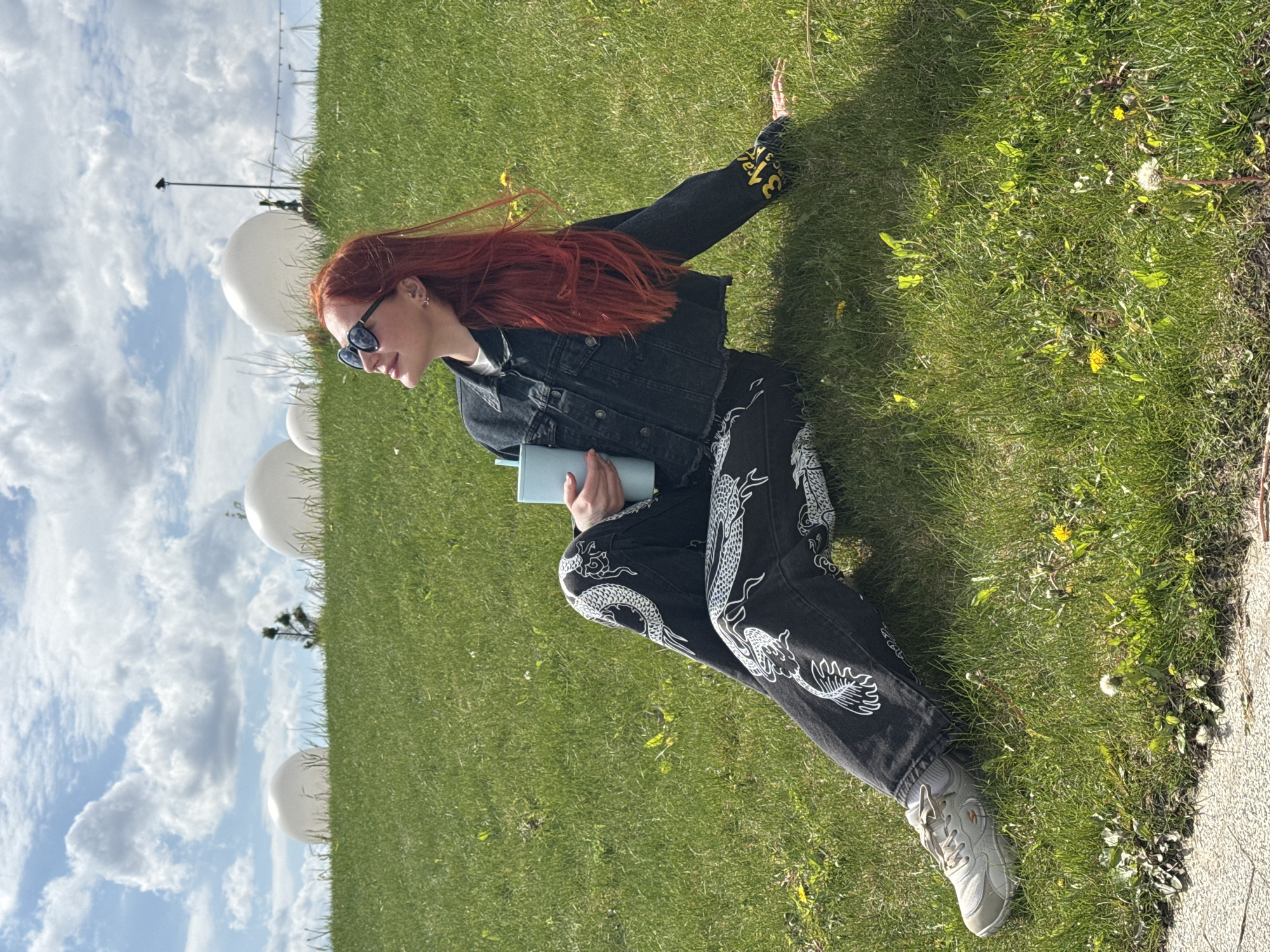 A young woman with red hair is sitting on a grassy hill. She is wearing sunglasses, a black denim jacket with yellow writing on the sleeve, black pants with white dragon designs, and gray sneakers. She is holding a light blue book or notebook. Behind her are several large, white spherical objects, possibly decorations or sculptures. The background includes a blue sky with fluffy white clouds. The location is Rostov-on-Don, Russia. The woman is resting on the hill, seemingly enjoying the outdoor environment.