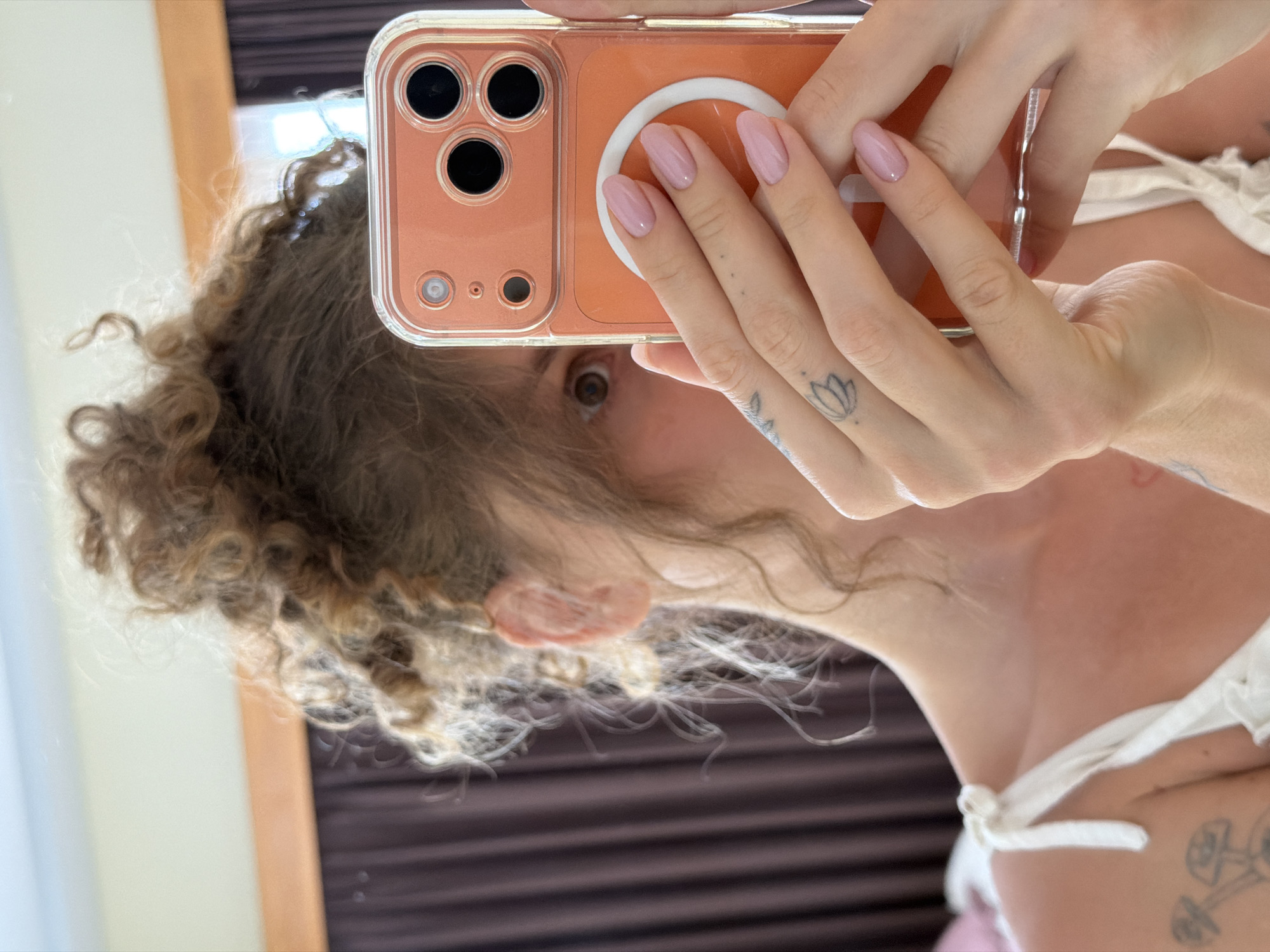A person with curly blonde hair and light skin is holding an orange iPhone to take a selfie. The phone partially obscures the person's face, but one brown eye is visible. The person is wearing a white top with straps. The background appears to be a mirror reflecting dark brown curtains or blinds. A tattoo is visible on the person's hand. Based on the information provided the image has no indications to place the image in North Kuta, Indonesia.