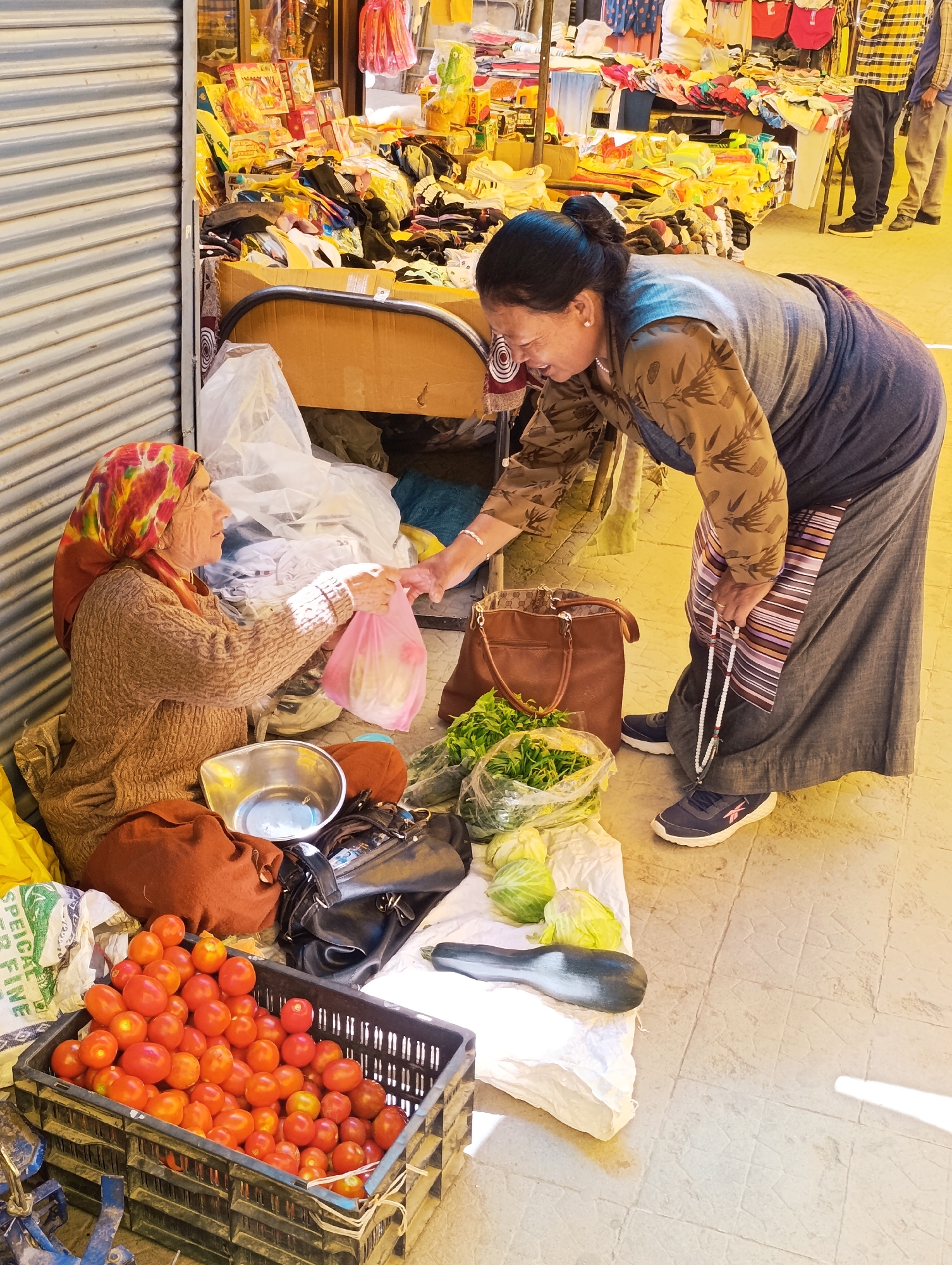 In Leh, India, a street vendor wearing a colorful headscarf and a brown knitted sweater sits on the ground by a stall. She is handing a pink plastic bag containing produce to another woman, who is dressed in a brown and patterned top and a long gray skirt. The second woman is leaning down and appears to be receiving the bag. Visible items include a crate overflowing with red tomatoes, bags of green vegetables such as lettuce and cabbage, and several handbags. The background reveals a market setting with various goods displayed on tables and racks, and a corrugated metal shutter.
