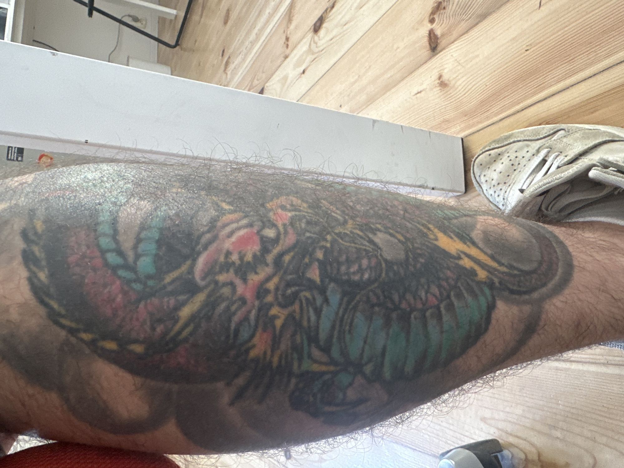 The image shows a close-up of a person's leg with a colorful dragon tattoo. The leg is covered in hair and is positioned on a wooden floor. A white shoe is visible in the lower right corner of the frame. To the right of the leg is a white object that appears to be part of a piece of furniture, possibly a door or a cabinet, which is positioned against a wall. The background shows a wooden floor with planks of varying shades of light brown. There is a white object in the background which may be a desk or a counter. The scene appears to be indoors. The image is a medium shot, focusing on the tattoo and the immediate surroundings. There is no explicit indication of the location being in Łódź, Poland.