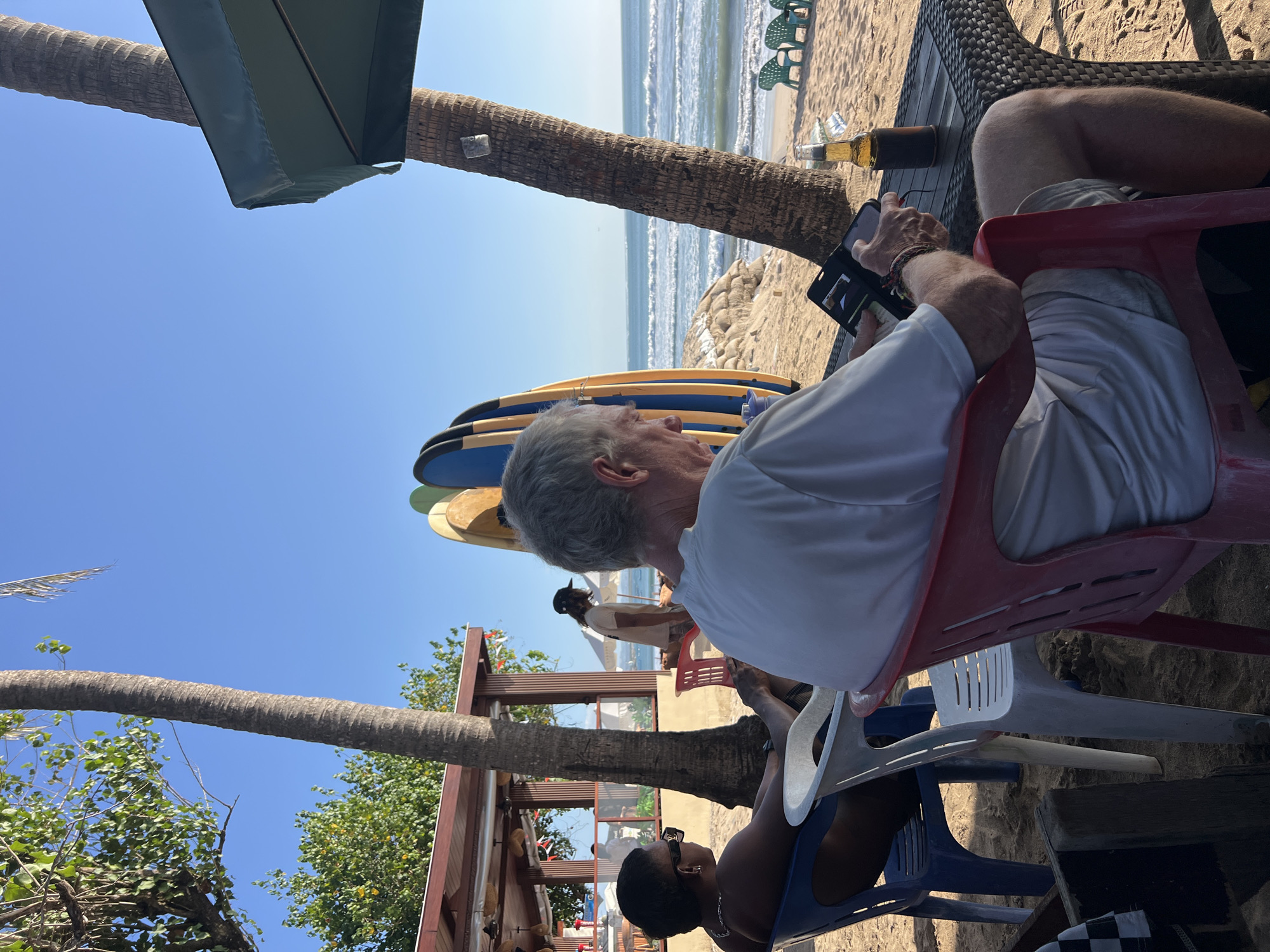 This image depicts a beach scene in Kuta, Indonesia, on a clear day. In the foreground, an older Caucasian man with gray hair is seated in a red chair, looking down at a black smartphone case he is holding in his hands. He is wearing a white t-shirt and light-colored shorts. To his left, a Black woman wearing sunglasses and a black top is seated in a blue chair. Next to the man, on a woven table, is a bottle of beer in a dark koozie and an ice-filled glass.
Behind the people, several surfboards are leaning against a palm tree. In the middle ground, a person wearing a tan shirt and a dark baseball cap walks towards the ocean. A beachfront structure with a red roof and open-air seating is visible to the left, with additional tables and chairs. The beach is sandy, with the ocean and gentle waves stretching to the horizon. Palm trees are scattered throughout the scene. In the background, several green beach chairs are arranged on the sand near the water.