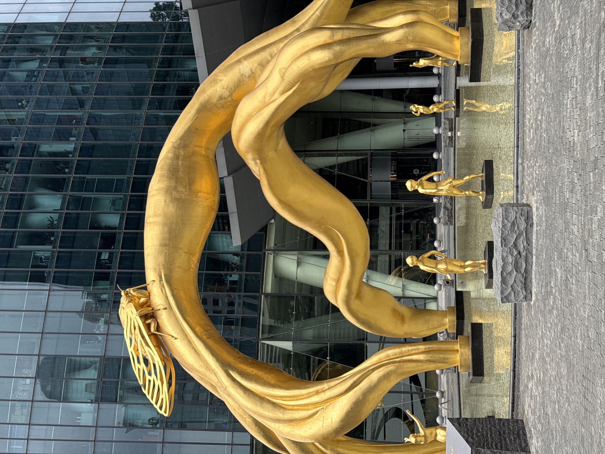 The image shows a large, golden, abstract sculpture in front of a modern, glass-walled building. The sculpture features flowing, ribbon-like forms. At the bottom left is a large, golden cicada sculpture attached to one of the ribbon-like forms. A series of small, golden figures stand on raised platforms, appearing to walk or run along a water feature. The paved area in the foreground suggests an urban environment. The Tianhe District, China location is implied through the architectural style and sculpture.