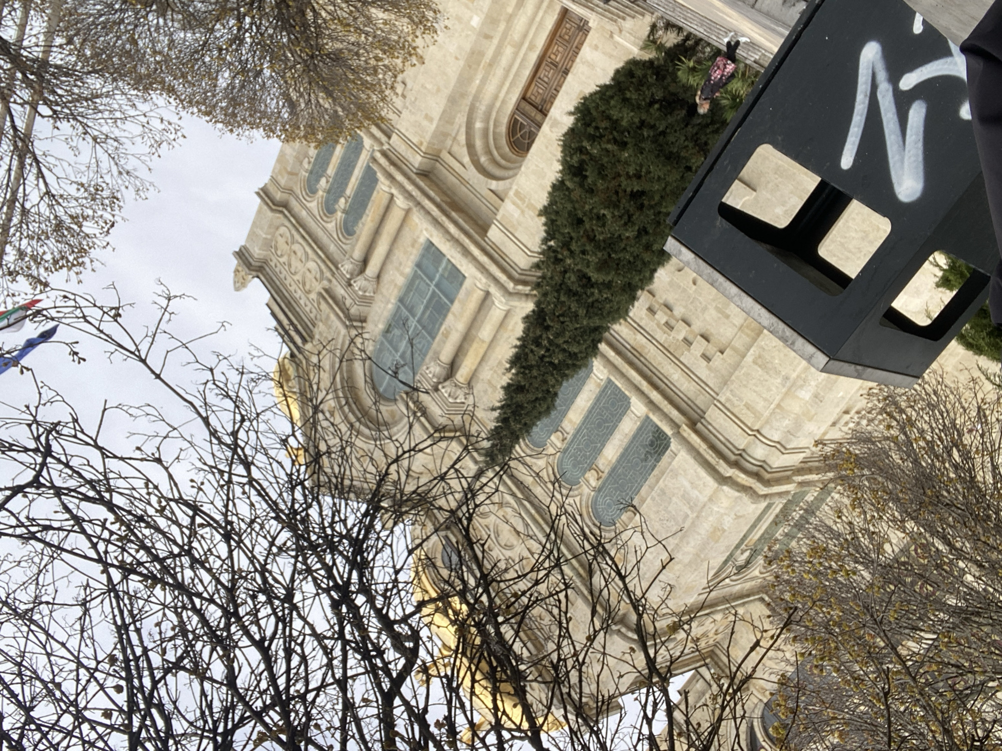 A low-angle shot shows the Cathedral of the Assumption of the Virgin in Varna, Bulgaria, interspersed with bare tree branches against a cloudy sky. The church is a light-colored stone with prominent arched windows and decorative detailing. A tall, narrow evergreen tree grows adjacent to the building. To the right, a dark, angular object with white graffiti-style lettering is in the foreground, partially obscuring the view. A person wearing a dark jacket and white shoes stands near the base of the tree, close to the church's wall.