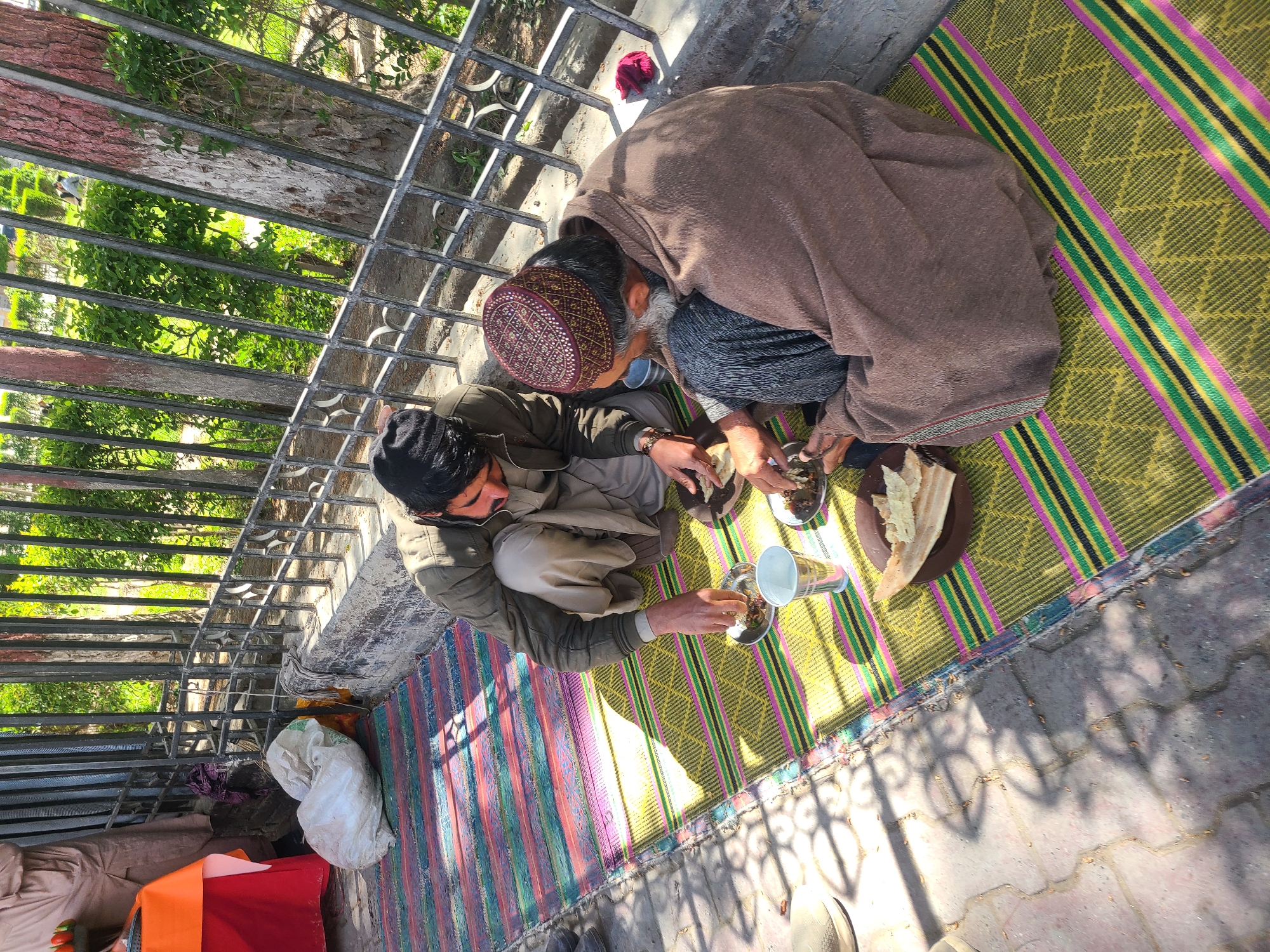 Two men sit on a woven mat outdoors in Quetta City Tehsil, Pakistan, sharing a meal. One man wears a brown overcoat and patterned cap. The other man wears an olive green jacket. They are surrounded by plates of food, a cup, and pieces of bread. A metal fence separates them from a tree and vegetation. The edge of the mat is visible along the brick pavement.
