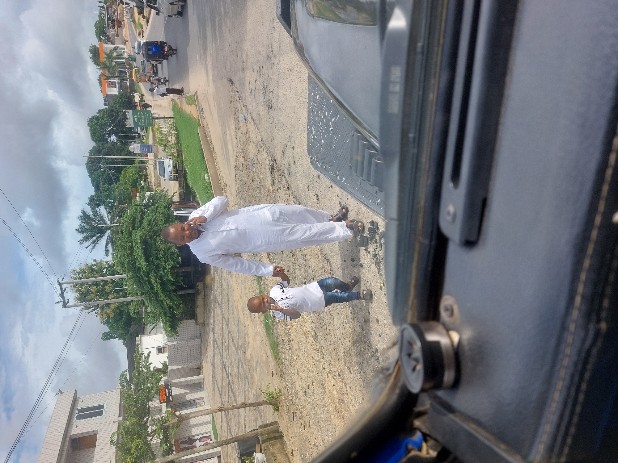 An outdoor shot depicts a man and child standing on a dirt and paved road in Dar es-Salaam, Tanzania. The man is wearing a long, white garment and brown sandals. He is holding the hand of a child dressed in a white button-down shirt, blue jeans, and brown shoes. The road has scattered debris. Buildings and greenery flank the road, along with cars and motorized vehicles in the background. The sky overhead is partly cloudy. The image is taken from inside a vehicle, with a partial view of the dashboard and window frame.