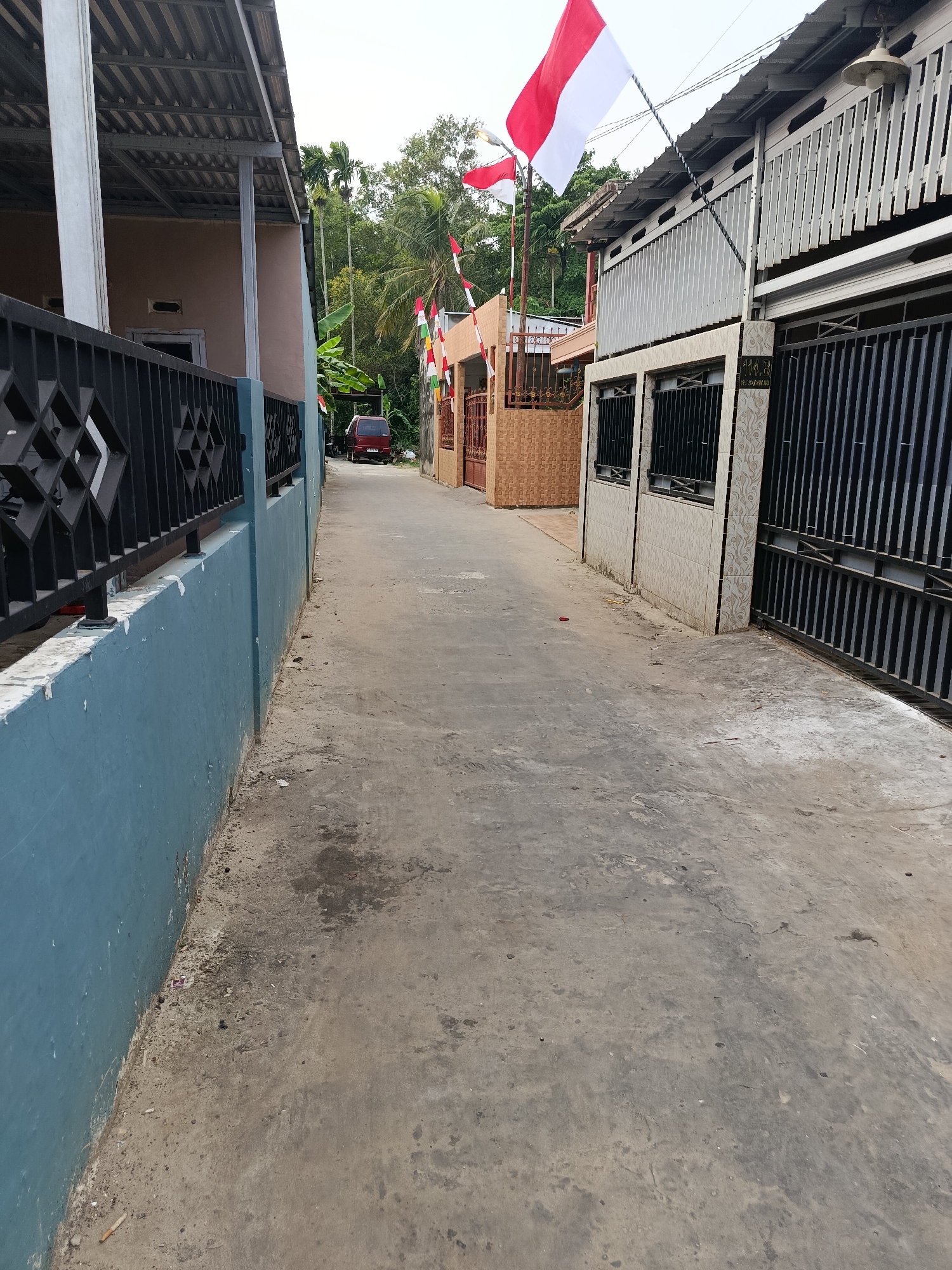 The image depicts a narrow, paved alleyway bordered by residential buildings and a concrete wall. Indonesian flags, red and white, are prominently displayed on poles along the right side of the alley, suggesting a celebration or national holiday. The buildings appear to be modest homes with metal roofs and fences. A red van is parked further down the alley, and lush greenery, likely trees and palms, are visible in the background. The overall impression is of a residential area in a tropical climate, possibly a neighborhood in Palembang, Indonesia, given the flags. The alleyway is clean but shows signs of wear on the pavement.