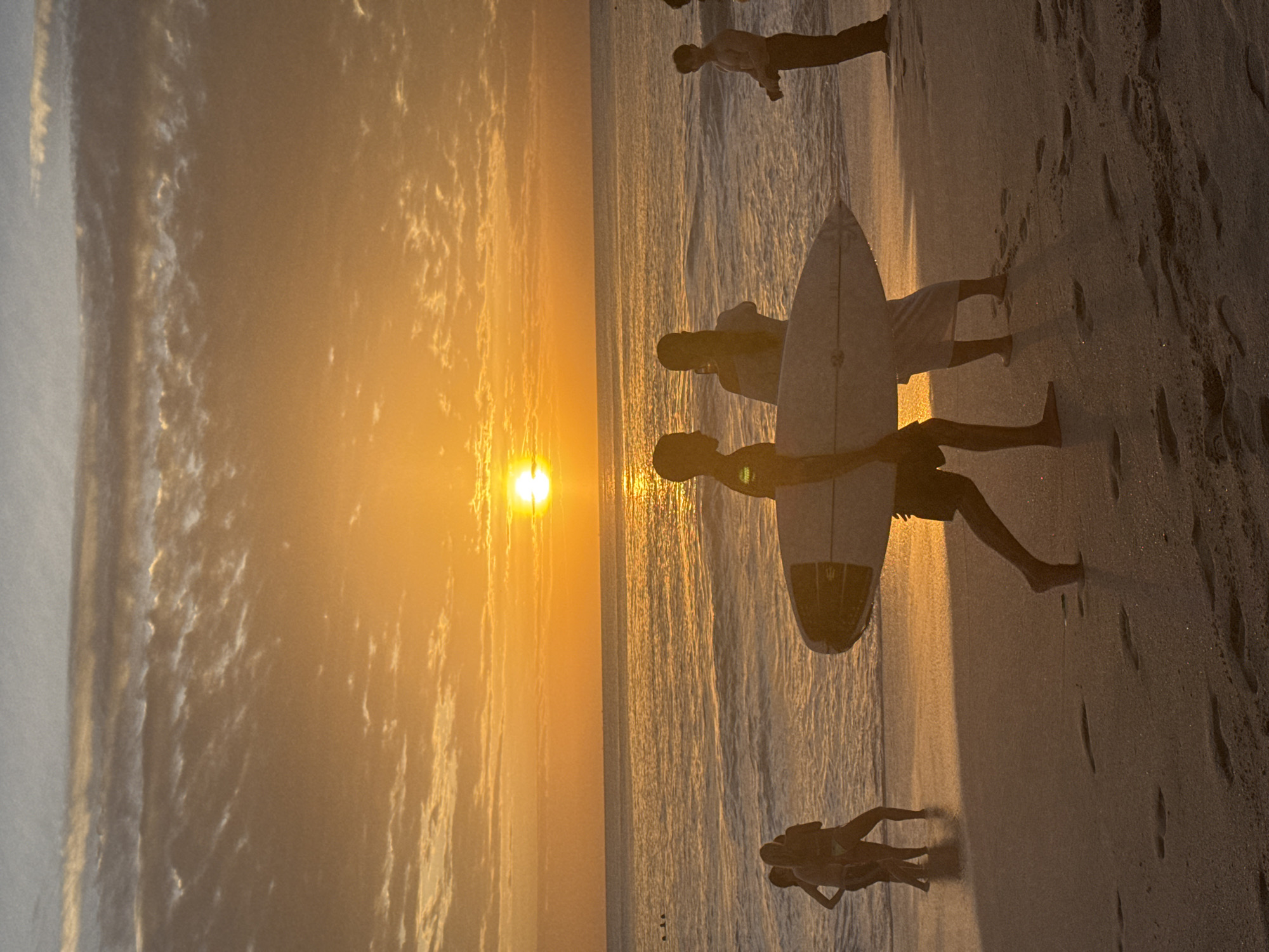 The image depicts a beach scene at sunset. The sun is low on the horizon, casting a warm orange glow across the sky and water. Clouds are visible in the sky, illuminated by the setting sun. The ocean has gentle waves and is reflecting the sunlight. Several people are present on the sandy beach. In the foreground, a person is walking with a surfboard. To their left, another person is carrying a surfboard, and to their left, two other figures stand together. Further out in the water, a few individuals are swimming. On the right side of the image, a person is standing, looking towards the water, and appears to be holding a camera or phone. The overall atmosphere is serene and captures the beauty of a sunset at the beach.