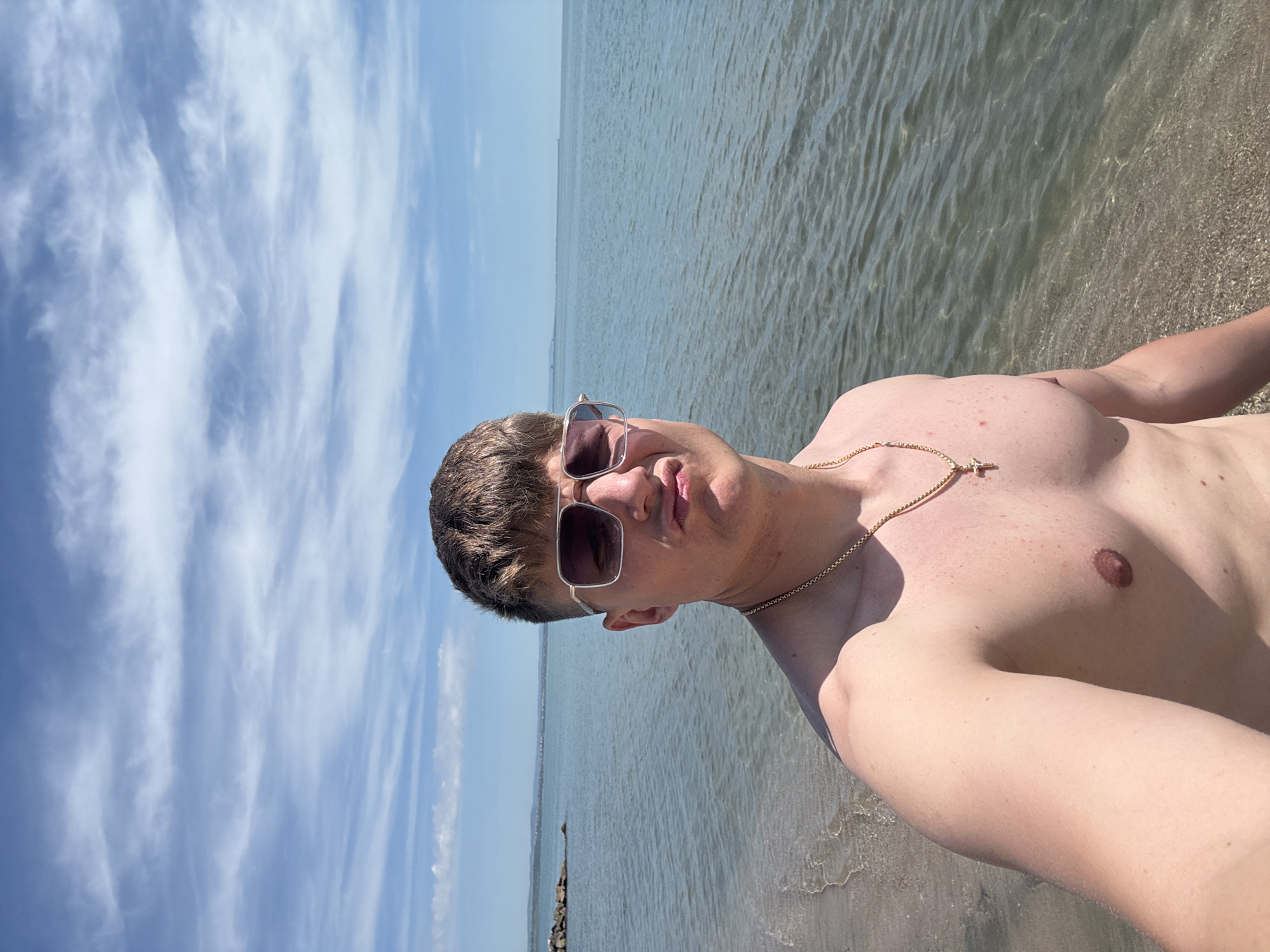 A man is standing on a beach, likely on the coast of Burgas, Bulgaria, as the background shows the sea and a cloudy sky. He is shirtless, wearing sunglasses, and a gold chain with a cross pendant. The water is calm and clear, indicating shallow depth near the shore. The beach appears sandy. A breakwater is slightly visible in the distance.