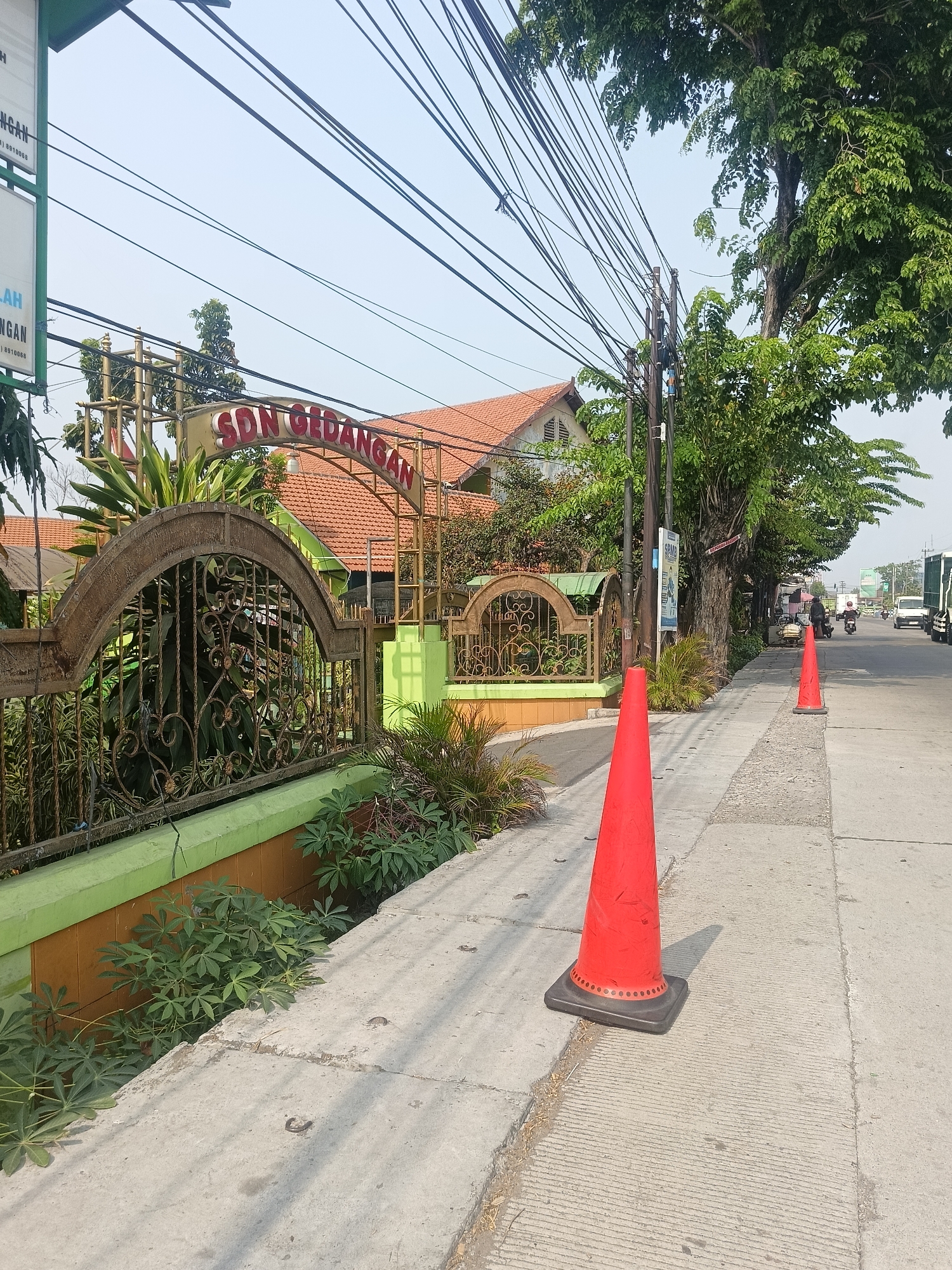 The image shows a street scene in Gedangan, Indonesia, with the entrance to SDN Gedangan visible. The school's entrance features an arched sign with red lettering that reads "SDN GEDANGAN" and is supported by a metal structure. In front of the entrance is an ornate metal fence and lush green plants. The street is paved, and a concrete sidewalk runs alongside it. Two bright orange traffic cones are placed on the sidewalk, with one closer to the foreground and another further down the street. In the background, trees with dense green foliage line the street, and utility wires are strung overhead. Vehicles, including a truck and motorcycles, are visible on the road, suggesting ongoing activity. The sky is clear and bright, indicating a sunny day.