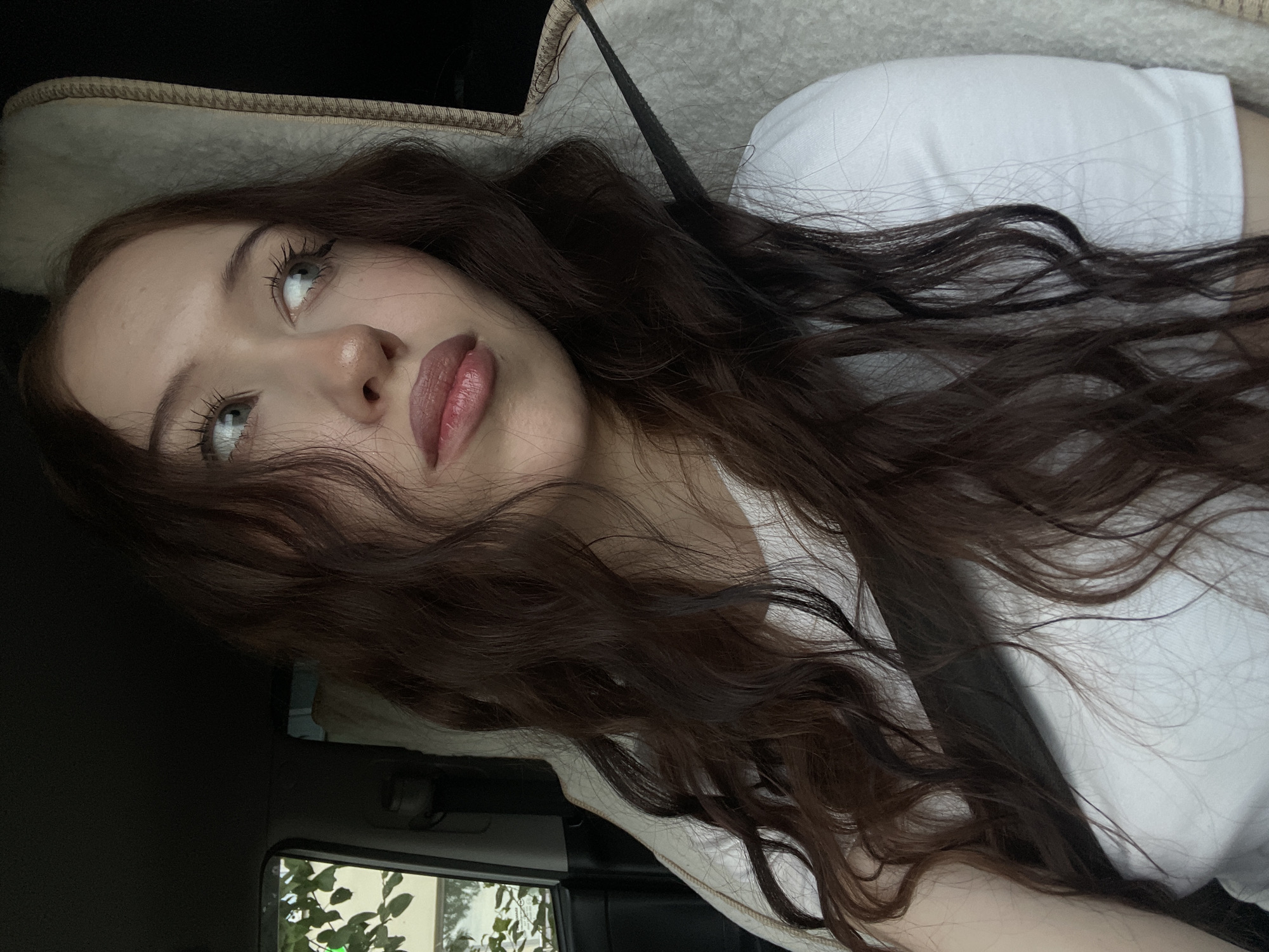 The image shows a young woman with long, dark brown, wavy hair sitting in the passenger seat of a car. She is wearing a white t-shirt and a seatbelt. Her head is turned to the side, and she is looking away from the camera with a neutral expression. The interior of the car is visible in the background, including a headrest and a window. Outside the window, greenery and trees can be seen, suggesting an outdoor location, possibly within a suburban or rural area of Votkinsk, Russia, given the context of the prompt. The lighting suggests it is daytime.