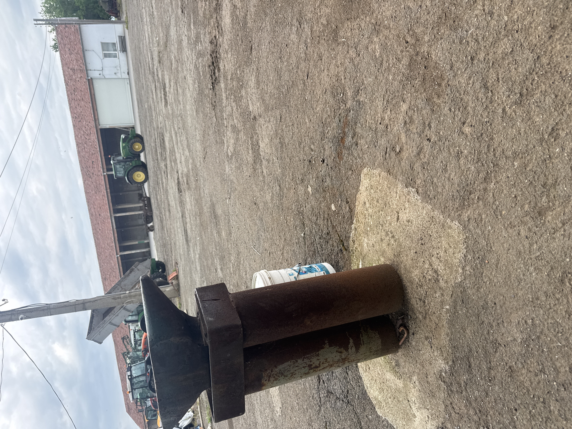 The image shows an anvil resting on two cylindrical metal pipes on a paved concrete surface. A white plastic bucket with blue and white graphics sits near the anvil. In the background, a large building with a red roof is visible, with several green and yellow tractors parked inside and around it. A utility pole and wires are present on the left. The location is likely near Sarnevo, Bulgaria, based on the presence of agricultural equipment and rural-style buildings. No people are visible.