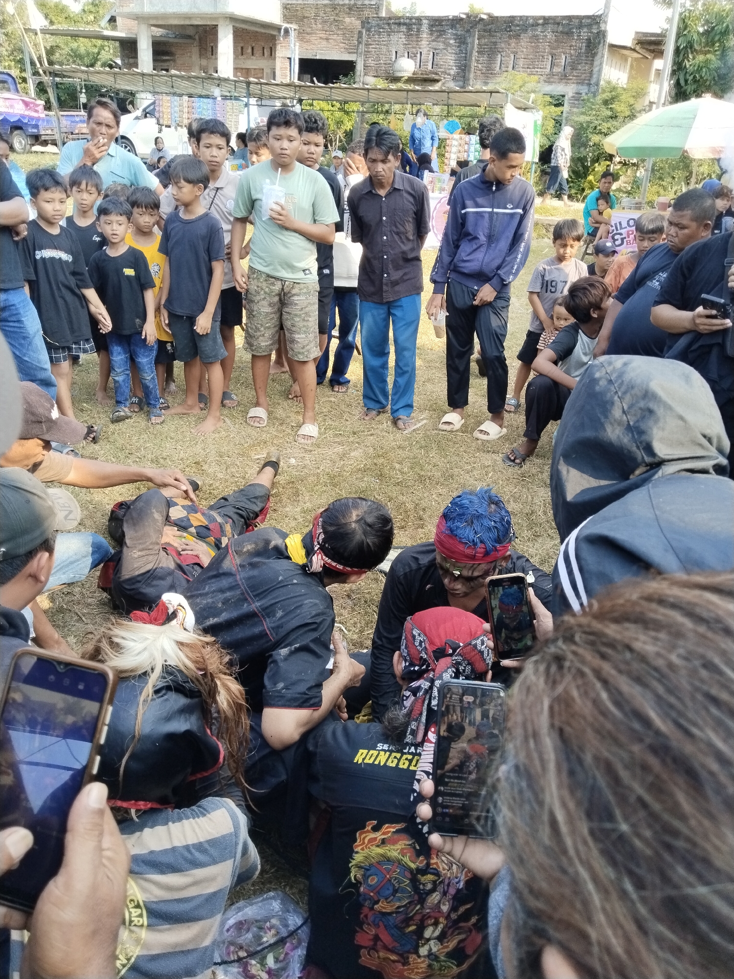 A crowd of people, including many children, has gathered in Kedawung, Indonesia, on a grassy area to witness a ritual or performance. The focus is on a group of individuals near the center of the frame who appear to be attending to a person lying on the ground. Several people are holding up cell phones, seemingly recording or capturing images of the event. The setting is a residential neighborhood, with houses and buildings visible in the background under a partly cloudy sky.