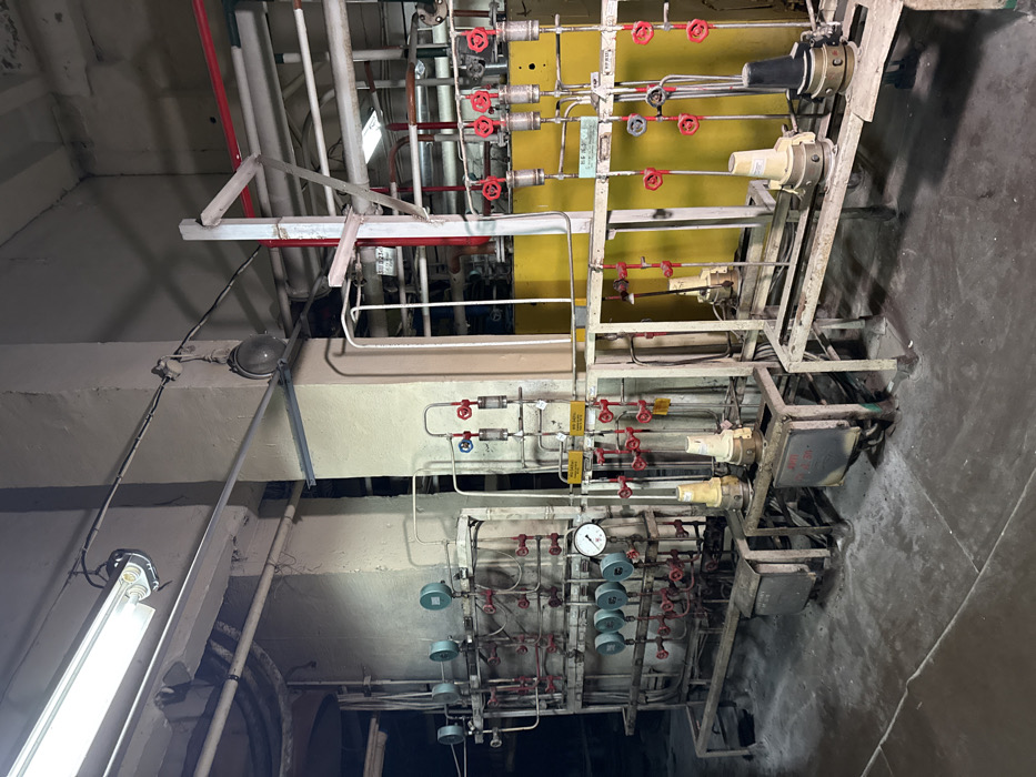 This image displays an interior view of an abandoned industrial facility, located in Чорнобиль, Ukraine, likely within the Chernobyl Exclusion Zone. The scene features a complex network of industrial process equipment, primarily composed of metal piping, numerous red-handled gate valves, and multiple circular pressure or flow gauges. These components are organized across two freestanding, multi-tiered metal racks. The left rack exhibits several gauges at varying heights, including one with a visible dial indicating a near-zero reading. The right rack features an array of vertical pipes equipped with individual red-handled valves and clear, tubular flow indicators. Behind the right-hand equipment, large yellow-painted panels or sections of machinery are visible. The structure of the room includes two prominent, light-colored concrete structural columns. Illumination is provided by a long, overhead fluorescent light fixture and several older, round industrial lamps mounted on the columns and ceiling. The concrete floor beneath the equipment appears dusty and worn. No individuals or active processes are present in the scene.