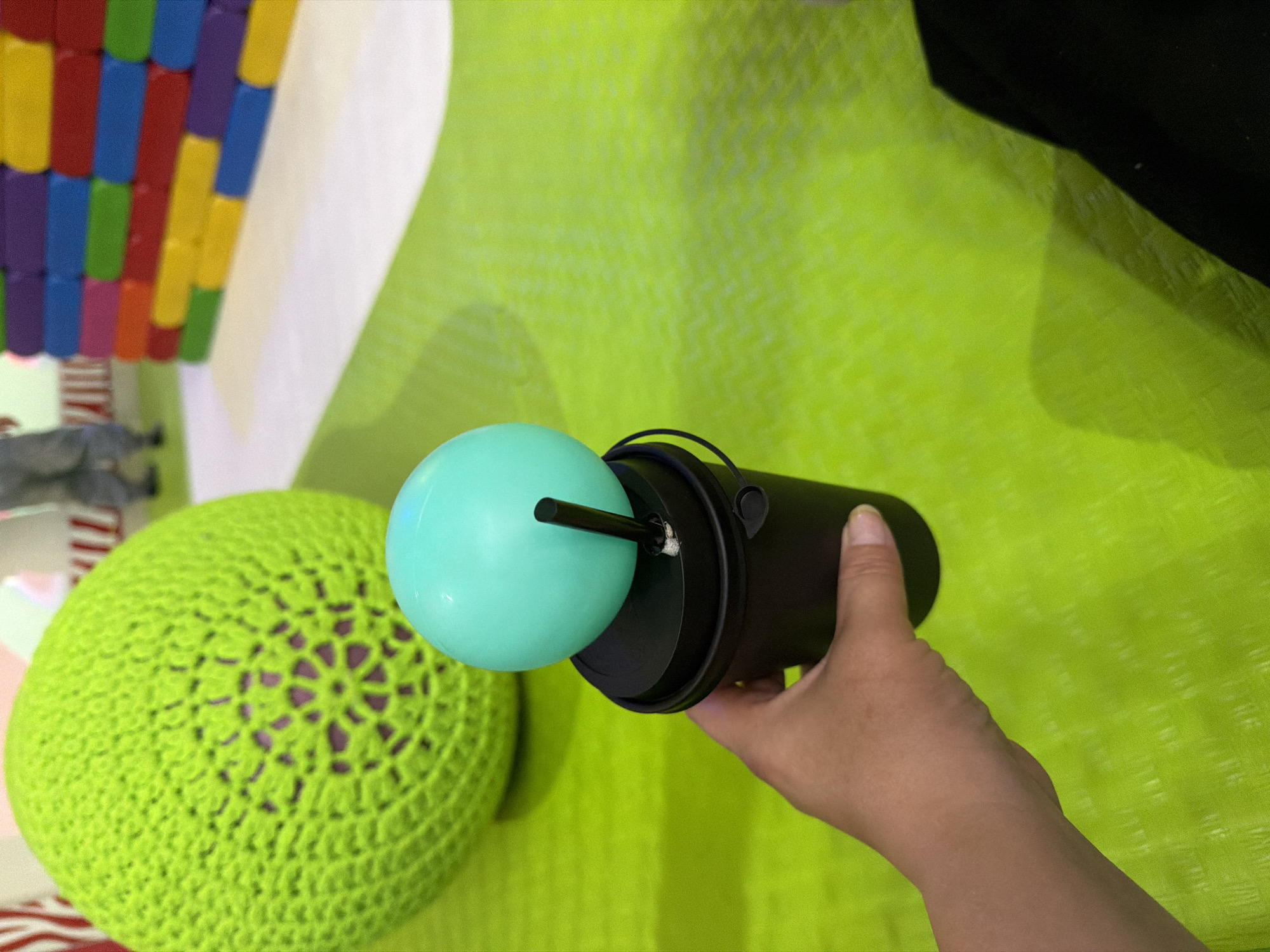 A person's hand holds a black cup with a light blue balloon on top and a straw poking through it. The cup is on a vibrant green surface with a wavy pattern. In the background, a large, textured, lime green ball is visible, and behind that, a wall made of colorful rectangular blocks is present. In the far background, the legs of a person wearing grey pants can be seen. The scene appears to be indoors in a brightly lit play area or entertainment venue.