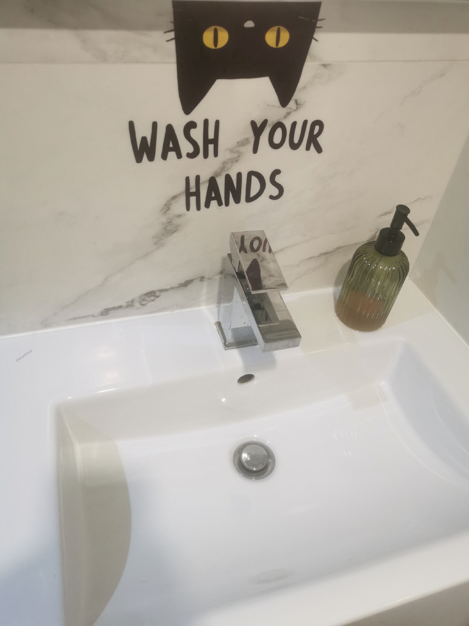 The image displays a modern bathroom sink area, photographed from an elevated perspective looking down. The background consists of a light-colored wall with grey veining, characteristic of marble or a marble-effect surface. Affixed to the wall is a black decal featuring the top portion of a cat's head with two prominent yellow, oval-shaped eyes. Below the cat's head, also in black decal, is the phrase "WASH YOUR HANDS" rendered in a bold, uppercase, handwritten-style font.
A white ceramic, rectangular sink with an integrated counter space dominates the lower portion of the frame. The word "FAYANS" is subtly visible in raised lettering on the upper left side of the sink basin. A contemporary chrome-plated single-lever faucet is mounted centrally on the sink's rear edge. Reflections on the faucet show distorted letters, possibly part of "SC DEMM". A circular chrome drain stopper is situated in the center of the sink basin. To the right of the faucet, a vertically ribbed, translucent green glass bottle with a black pump dispenser contains an amber-colored liquid, presumably liquid soap.
No individuals are present in the scene, and no actions or interactions are depicted. The environment appears clean and well-lit. There is no visual information within the image to determine its location in the city of Blagoevgrad, Bulgaria.
