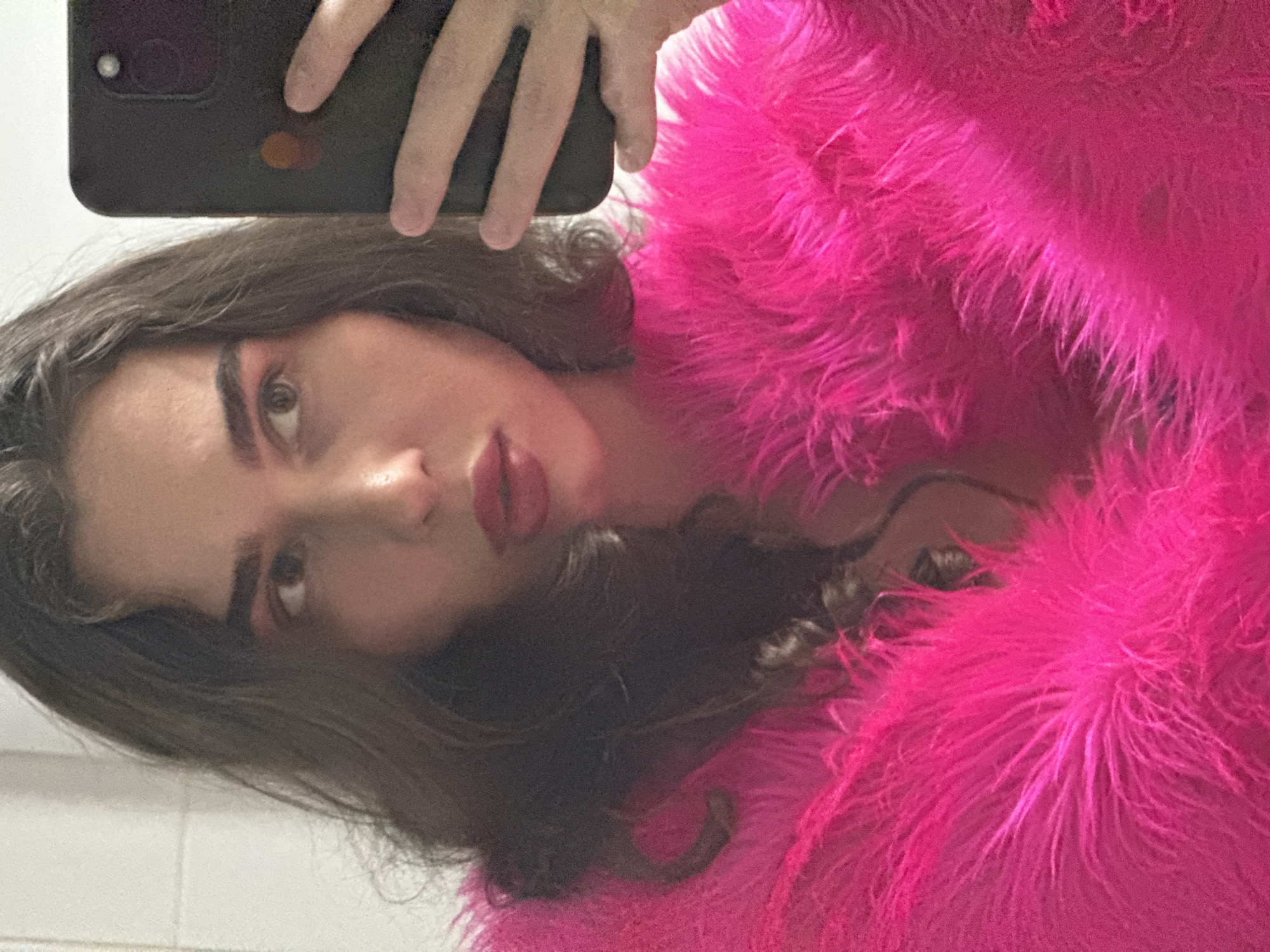 A woman is taking a selfie in a bathroom. She is wearing a bright pink fluffy jacket and has dark, wavy hair. Her makeup includes pink eyeshadow and lipstick. She is holding a black smartphone in her right hand. The background appears to be a tiled wall. The image does not contain information indicating the location is the city of Kazan, Russia.