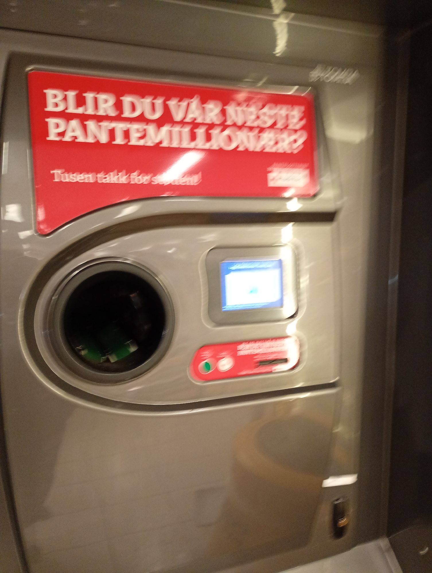 The image shows a close-up of a reverse vending machine, likely in a public space. The machine is gray with a red panel displaying text in white sans-serif font. The text reads "BLIR DU VÅR NESTE PANTEMILLIONÆR?" which translates to "WILL YOU BE OUR NEXT BOTTLE MILLIONAIRE?". Below this, in smaller font, it says "Tusen takk for støtten!" which means "Thank you for your support!". A circular opening where bottles are inserted is visible on the left. To the right of the opening, there is a small screen displaying a light blue and white interface. Below the screen is a slot for inserting items, with red and green buttons on either side. The machine appears to be made of plastic and metal. The background is out of focus, but suggests an indoor environment. The lighting is artificial and casts some reflections on the machine's surface. There are no people visible in the image.