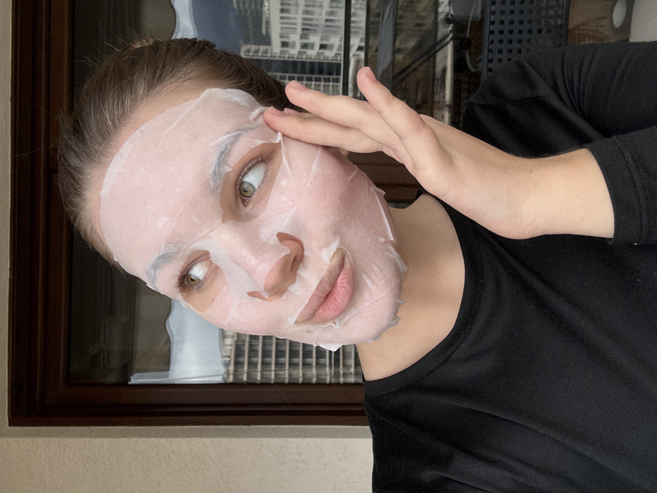 A female individual is depicted in a close-up, chest-up shot, positioned on what appears to be a balcony or terrace in Budva, Montenegro. She has light-toned skin, green eyes, and dark hair pulled back, with strands visible around the forehead and temples. Her face is covered with a translucent white sheet mask, which has cutouts for her eyes and mouth. Her lips are slightly parted. She is wearing a plain black long-sleeved top with a rounded neckline. Her right hand is raised, with fingers gently touching the left side of the sheet mask near her temple. She is looking upwards and to her right.
The background shows a light-colored wall to the left. Behind her, a large, dark-framed window or sliding door reflects and reveals a cityscape. Through the window, multiple high-rise buildings are visible, some appearing fully constructed, others with visible structural elements suggesting ongoing construction. Distant mountains are present under an overcast sky, characteristic of the coastal mountainous region of Budva.